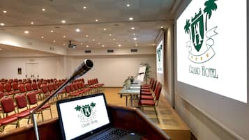 Une salle de conférence avec des rangées de chaises rouges, un pupitre et un ordinateur portable affichant le logo du Grand Hôtel.
