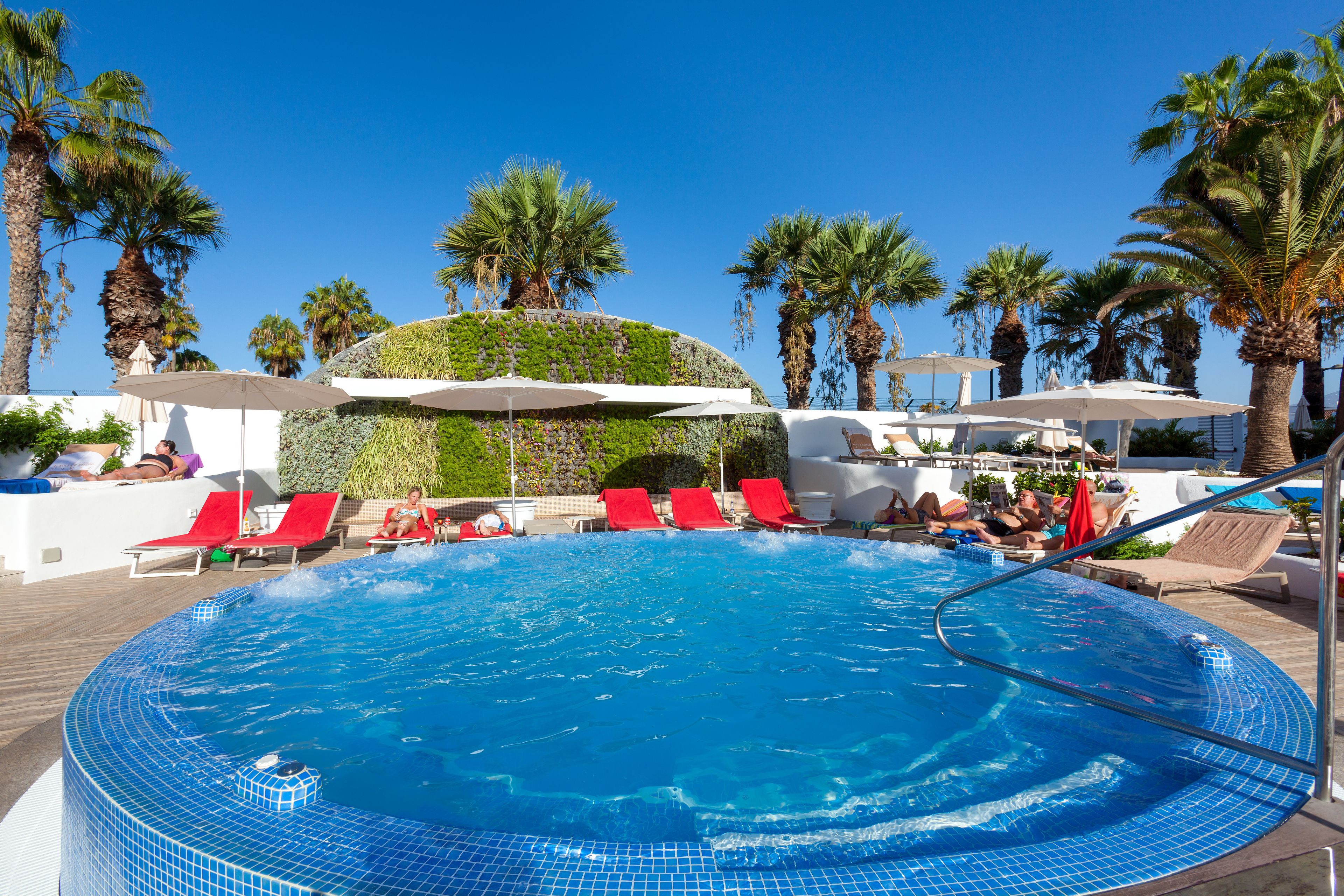Una piscina con sillas reclinables rojas y sombrillas, rodeada de palmeras y jardines.