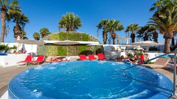 Una piscina con sillas reclinables rojas y sombrillas, rodeada de palmeras y jardines.
