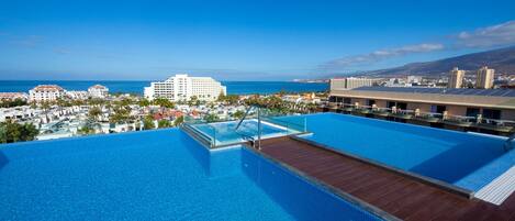 A rooftop infinity pool with a wooden deck and glass balustrade, overlooking a coastal cityscape and the ocean.