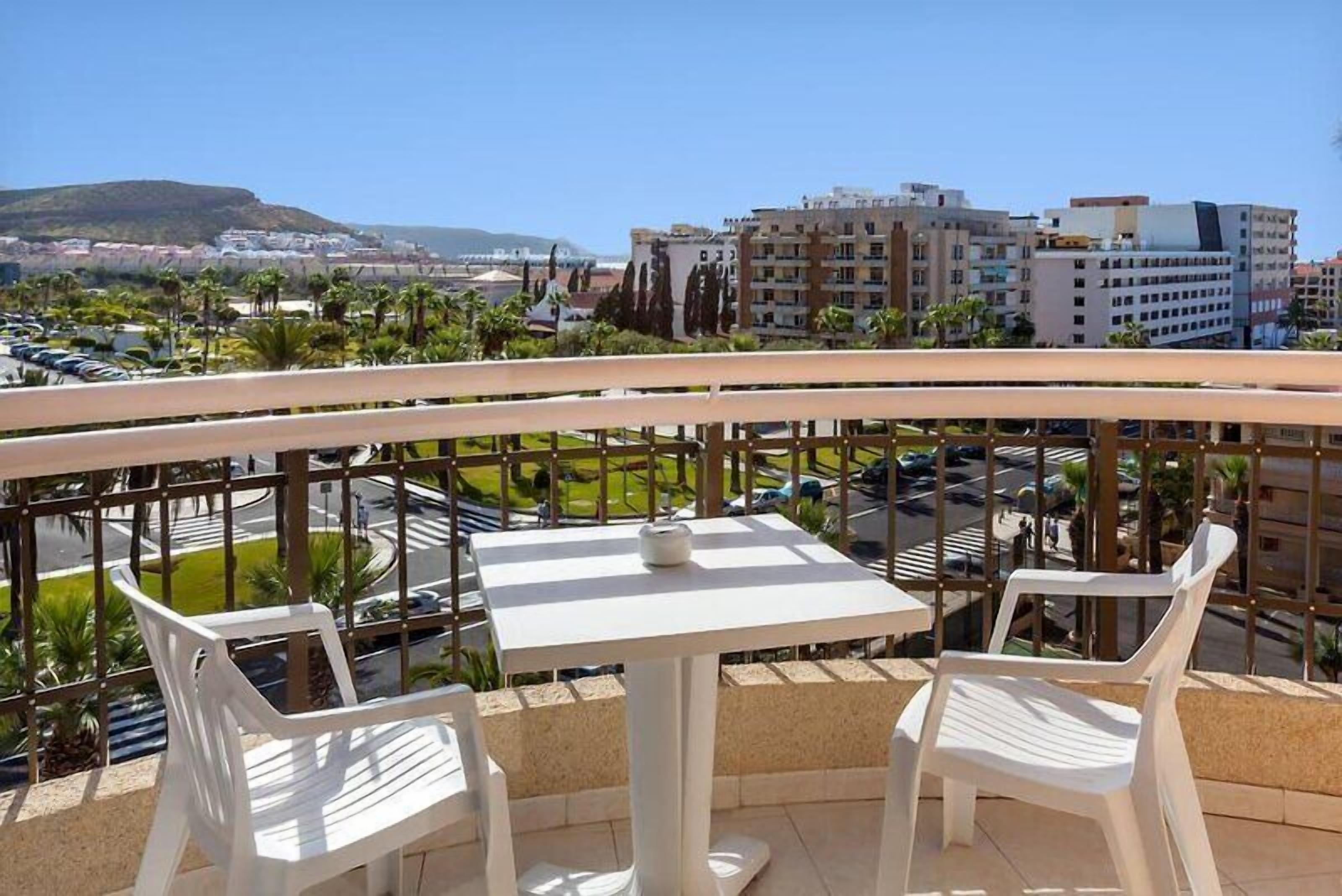 A balcony with a table and chairs, overlooking a cityscape with buildings and palm trees.