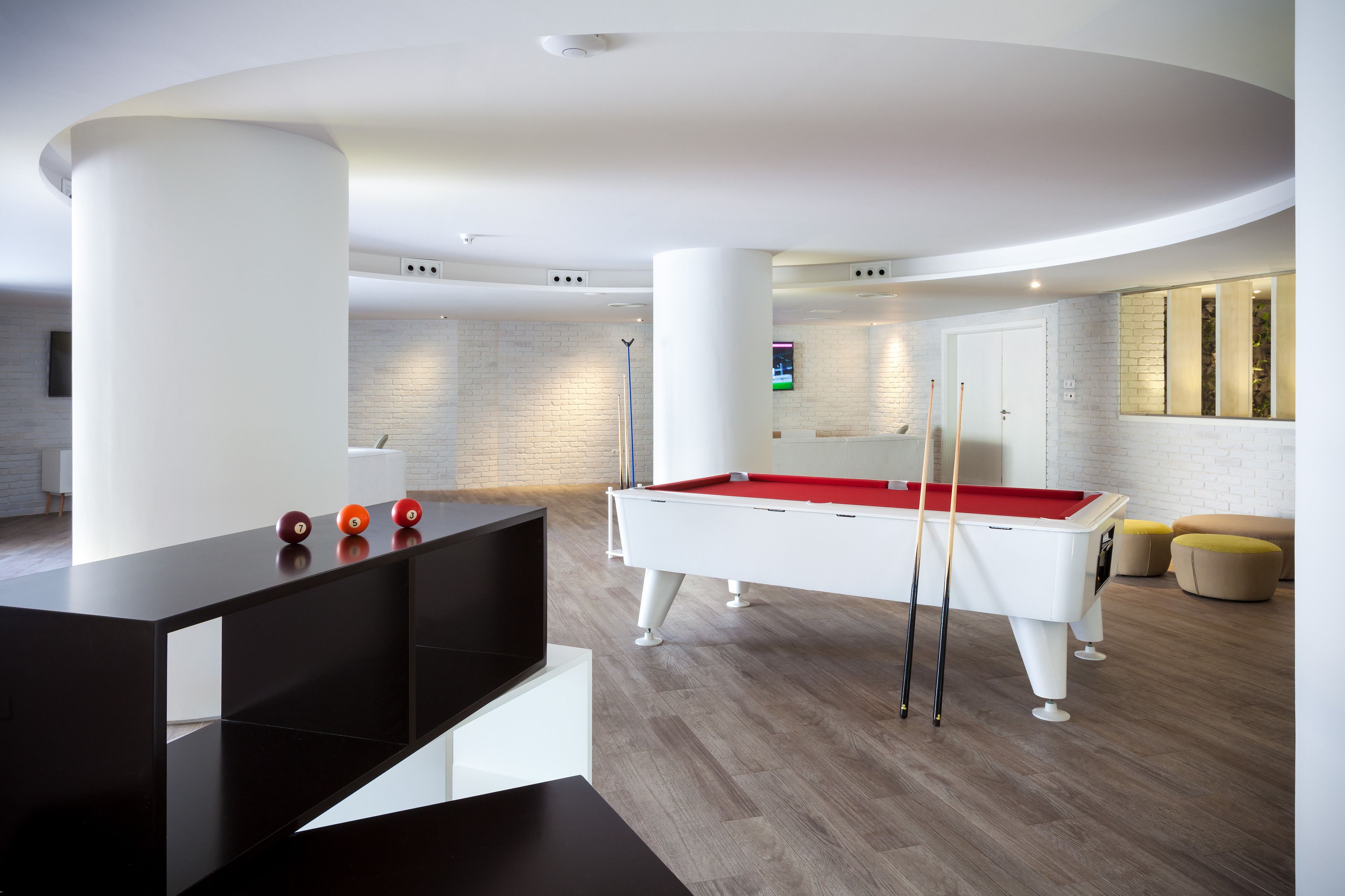 A modern pool table in a well-lit room with a minimalist design, featuring a white pool table, red felt, and a black pool cue rack.