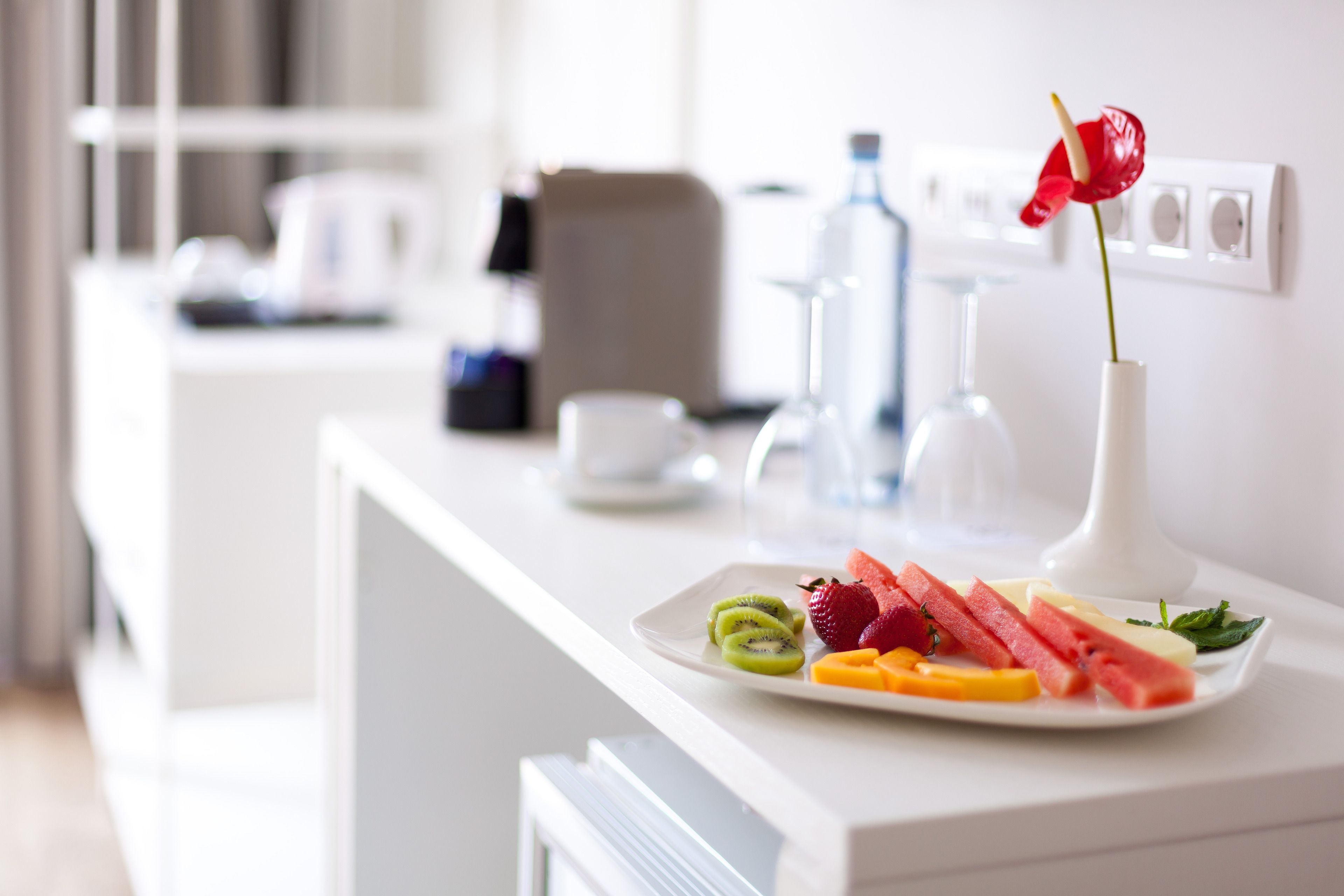 Una mesa de desayuno con un plato de fruta y un jarrón con una flor roja.