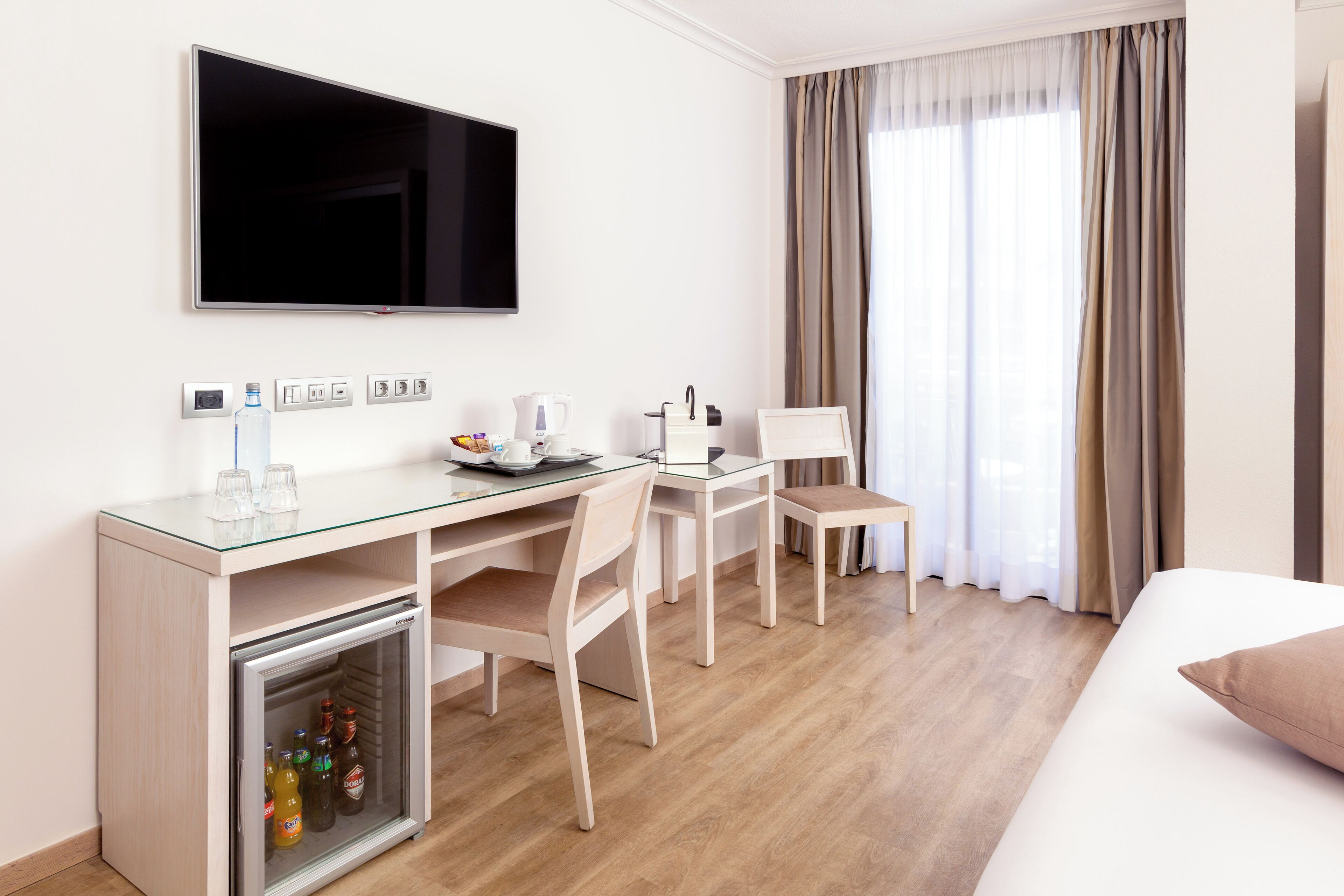 Habitación de hotel con cama, escritorio con televisor, refrigerador minibar y ventana con cortinas.