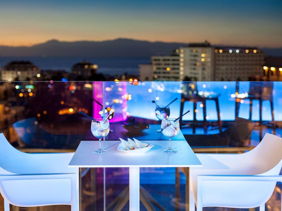 A rooftop terrace with a table and chairs, overlooking a cityscape and mountains.