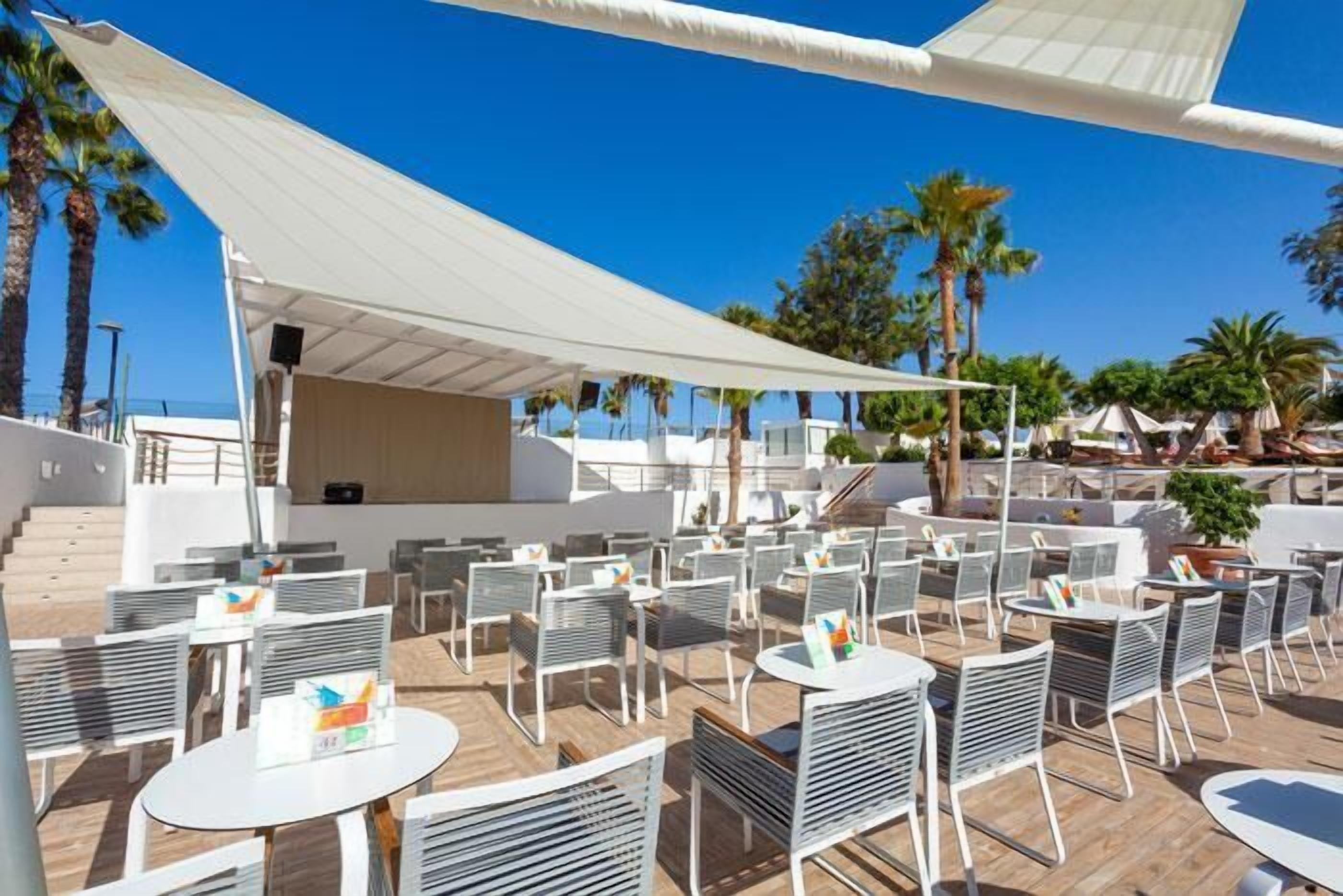 An outdoor seating area with tables and chairs, shaded by a large white canopy, surrounded by palm trees and a clear blue sky.