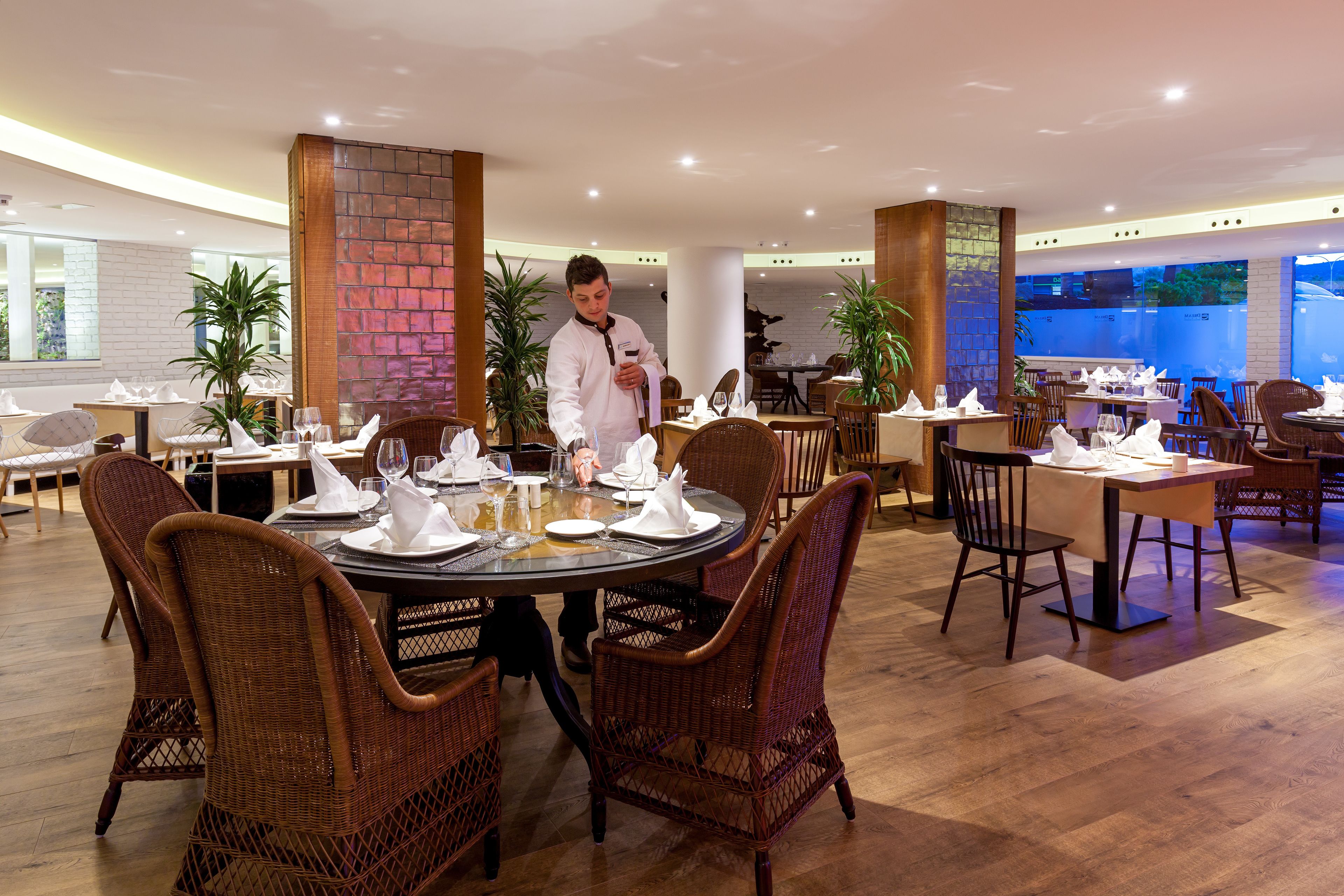 A restaurant with round tables, wicker chairs, and a waiter serving.