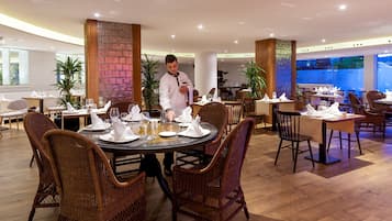 A restaurant with round tables, wicker chairs, and a waiter serving.