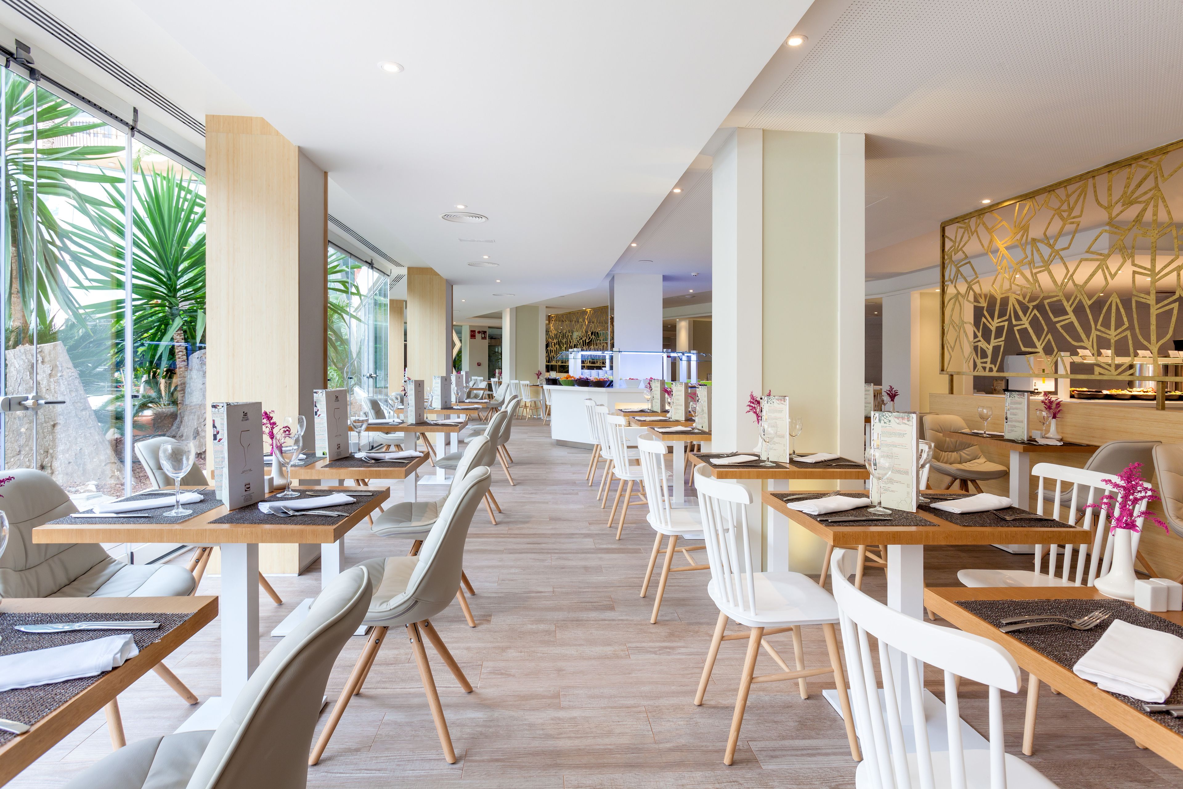 A modern restaurant with long tables and white chairs, large windows with plants, and a decorative wall on the right.