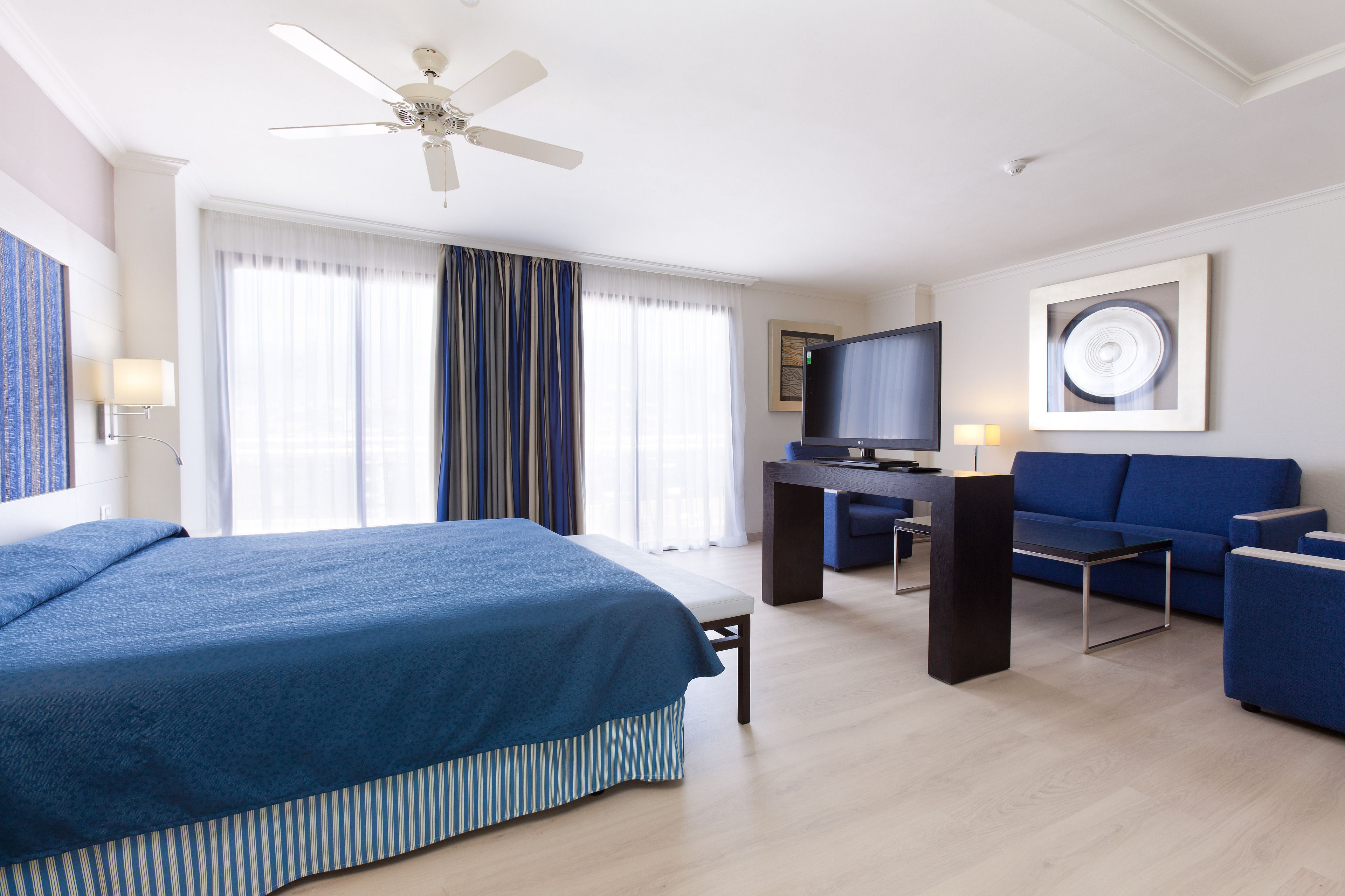Een hotelkamer met een groot bed, een bureau met een televisie, een bank en een plafondventilator.