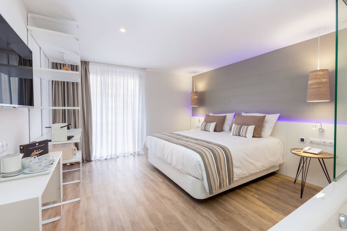 A modern hotel room with a large bed, a desk with a flat-screen TV, a wardrobe, and a bedside table with a lamp.