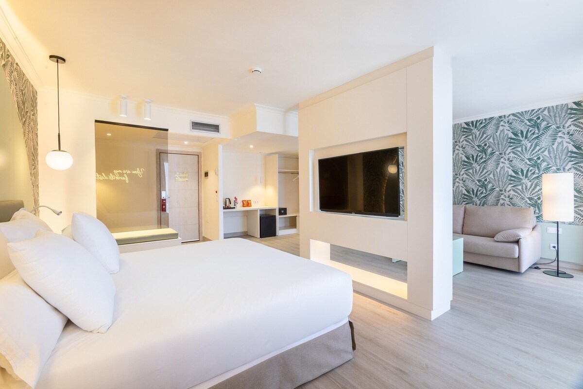 A modern hotel room with a large bed, a flat-screen TV mounted on the wall, a sofa, and a desk area.