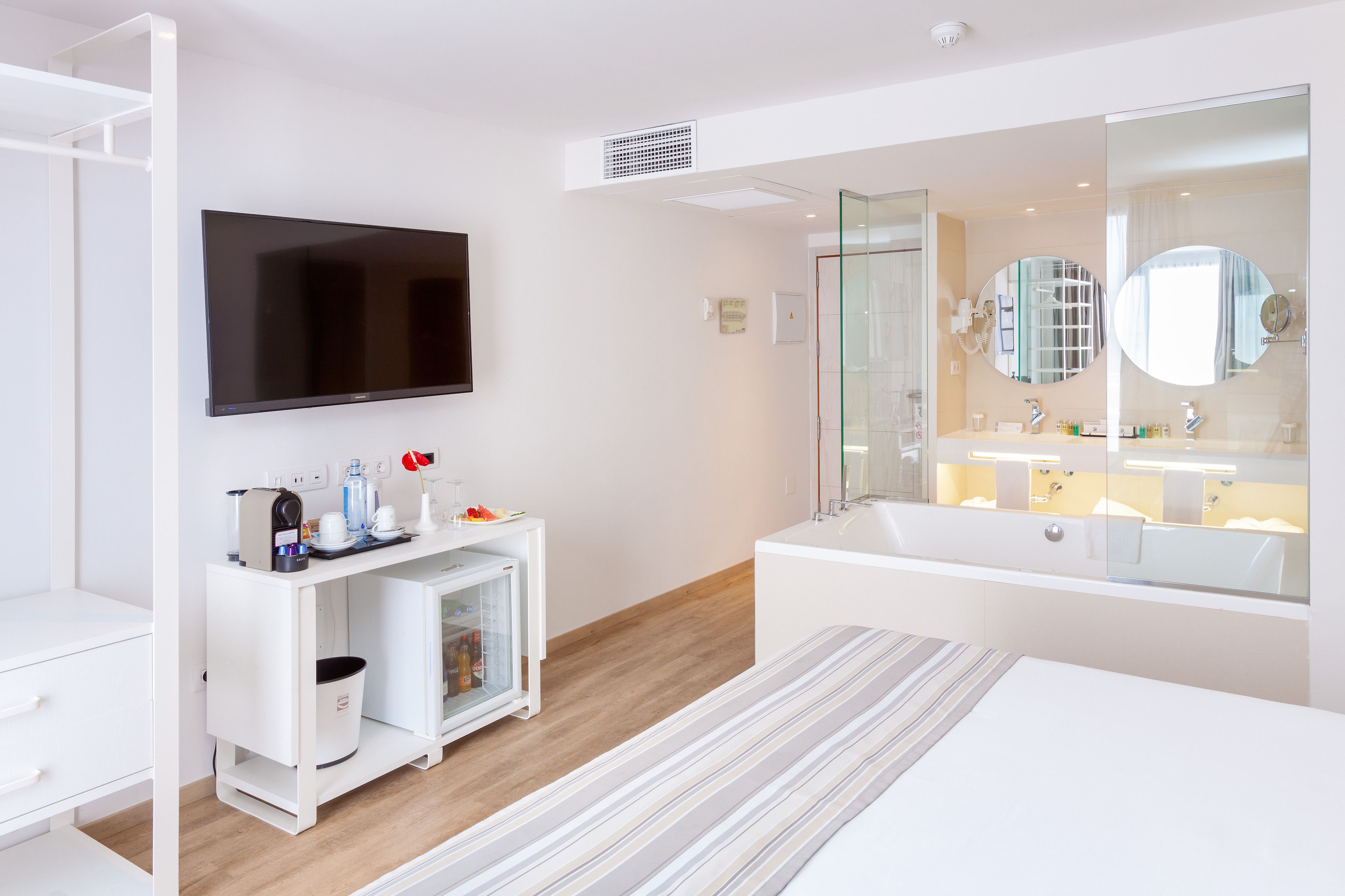 Una habitación de hotel moderna con una televisión de pantalla plana, un escritorio blanco con una cafetera, un mini refrigerador, un espejo grande, una bañera y un tapete a rayas.