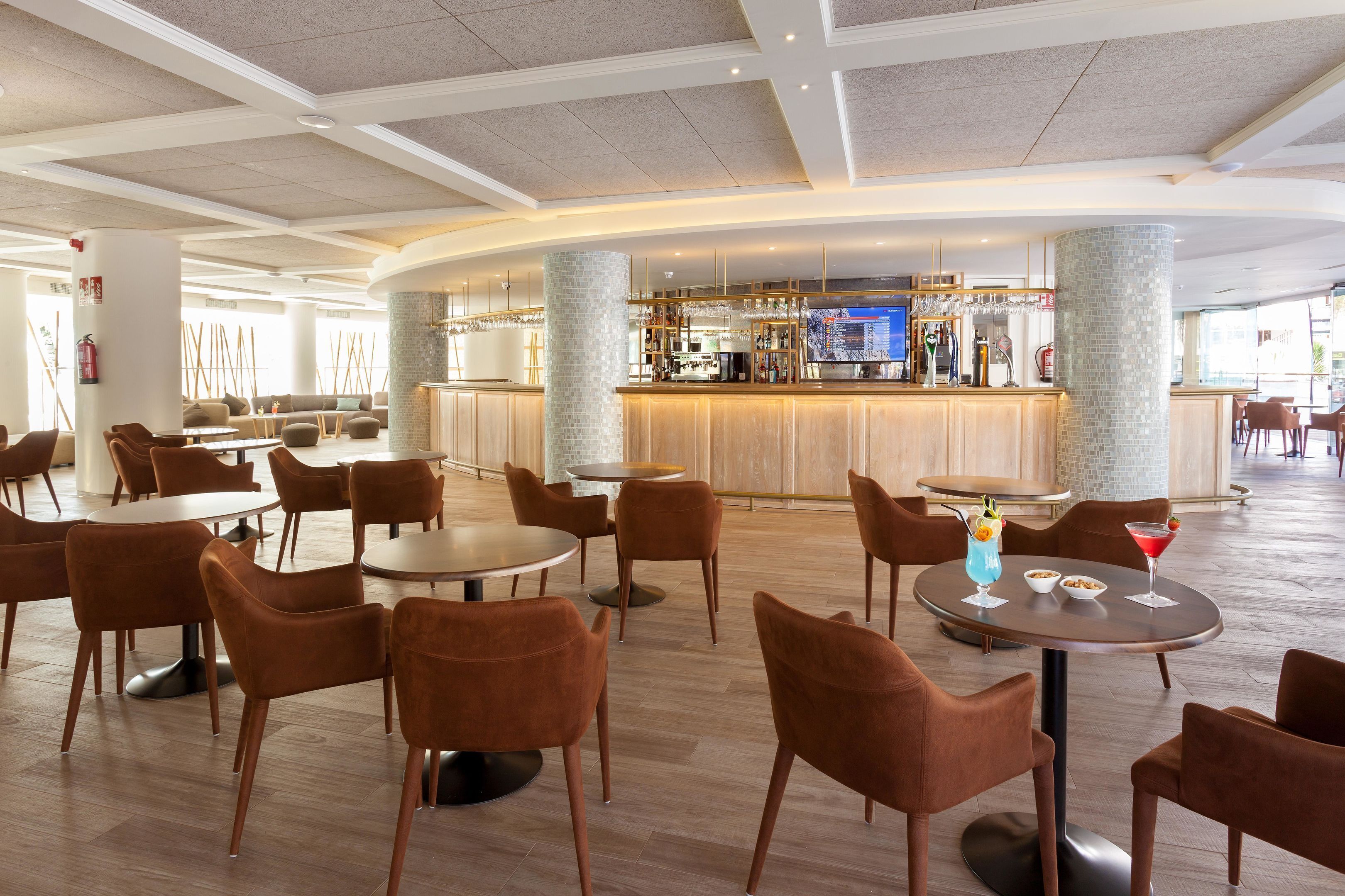 Eine moderne Hotellobby mit runden Tischen und braunen Stühlen, ein Barbereich mit Theke und Barhocker sowie eine große Säule.