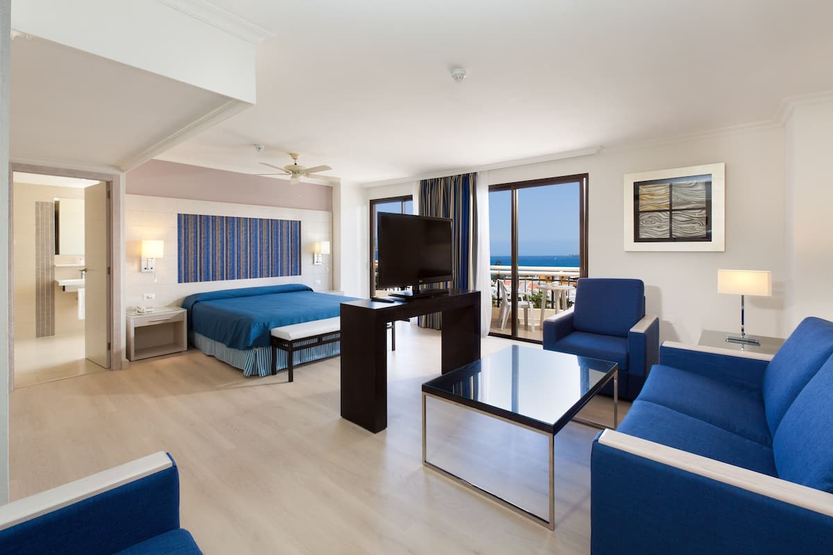 A modern hotel room with a balcony, a bed, a desk, and a seating area.