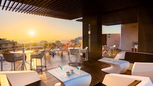 A rooftop lounge with white furniture, a wooden deck, and a sunset view.