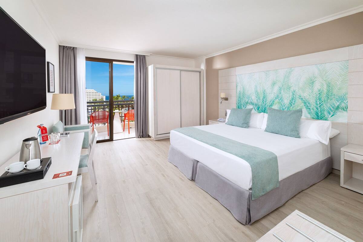 A hotel room with a bed, a desk with a coffee maker, a TV, and a balcony with a view of the city.
