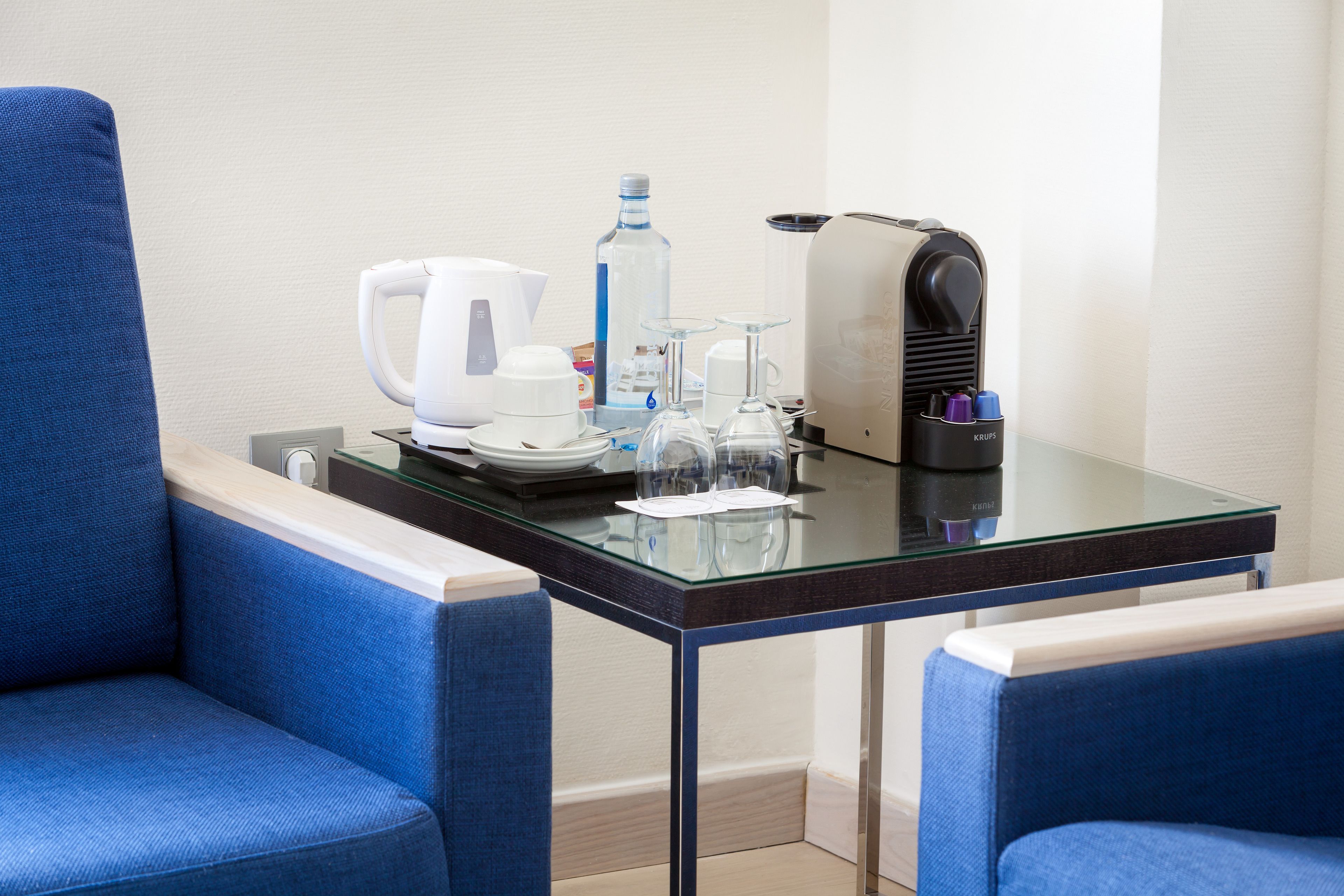 Habitación de hotel con una mesita de vidrio, una tetera, una botella de agua, una cafetera y un tostador. Hay dos sillones tapizados en azul.