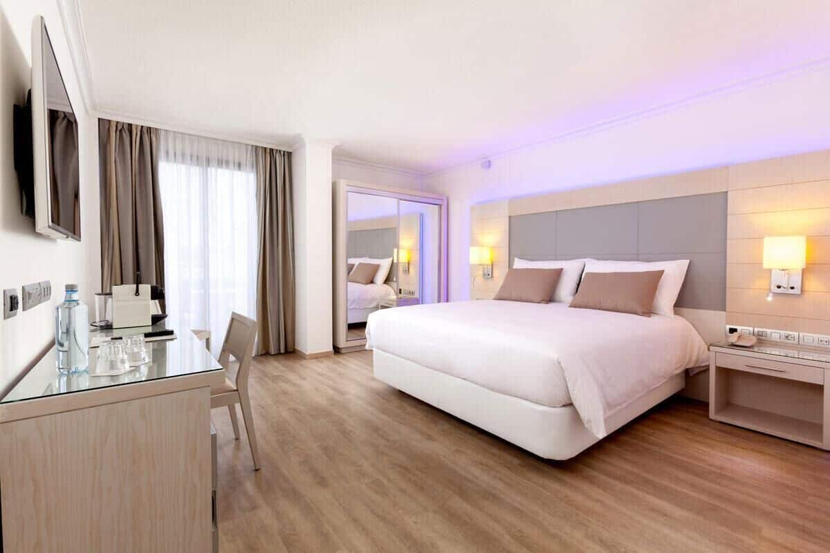 A hotel room with a large bed, bedside tables, a desk with a chair, a TV, and a mirror.