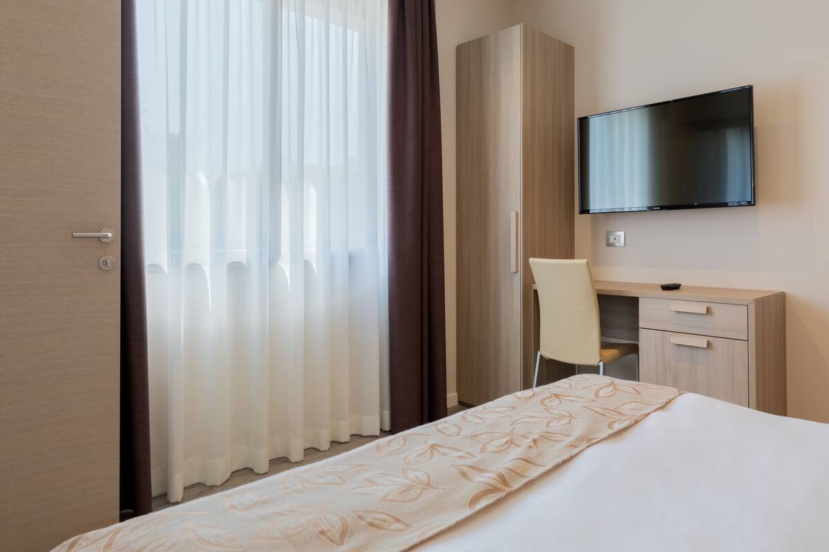 A hotel room with a bed, a desk, a chair, a television, and a wardrobe.