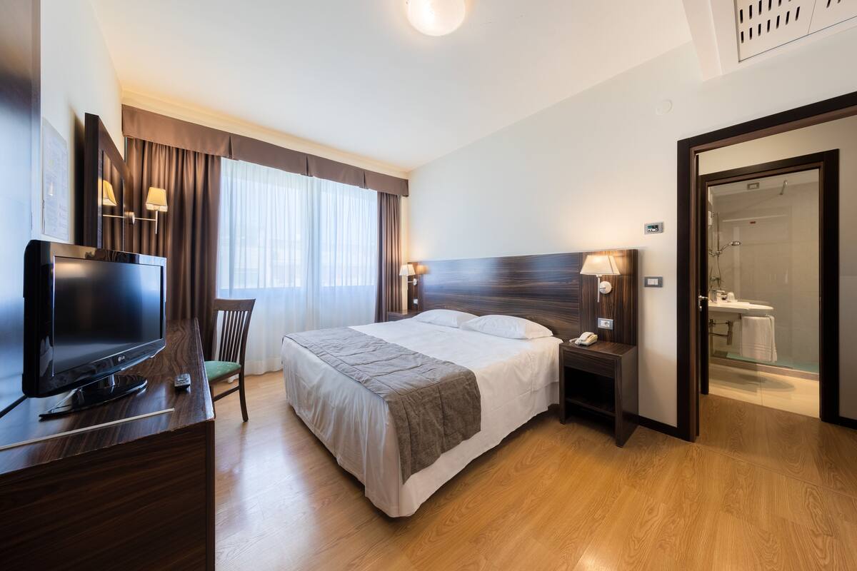 A hotel room with a bed, a TV, a desk, and a bathroom visible through an open door.