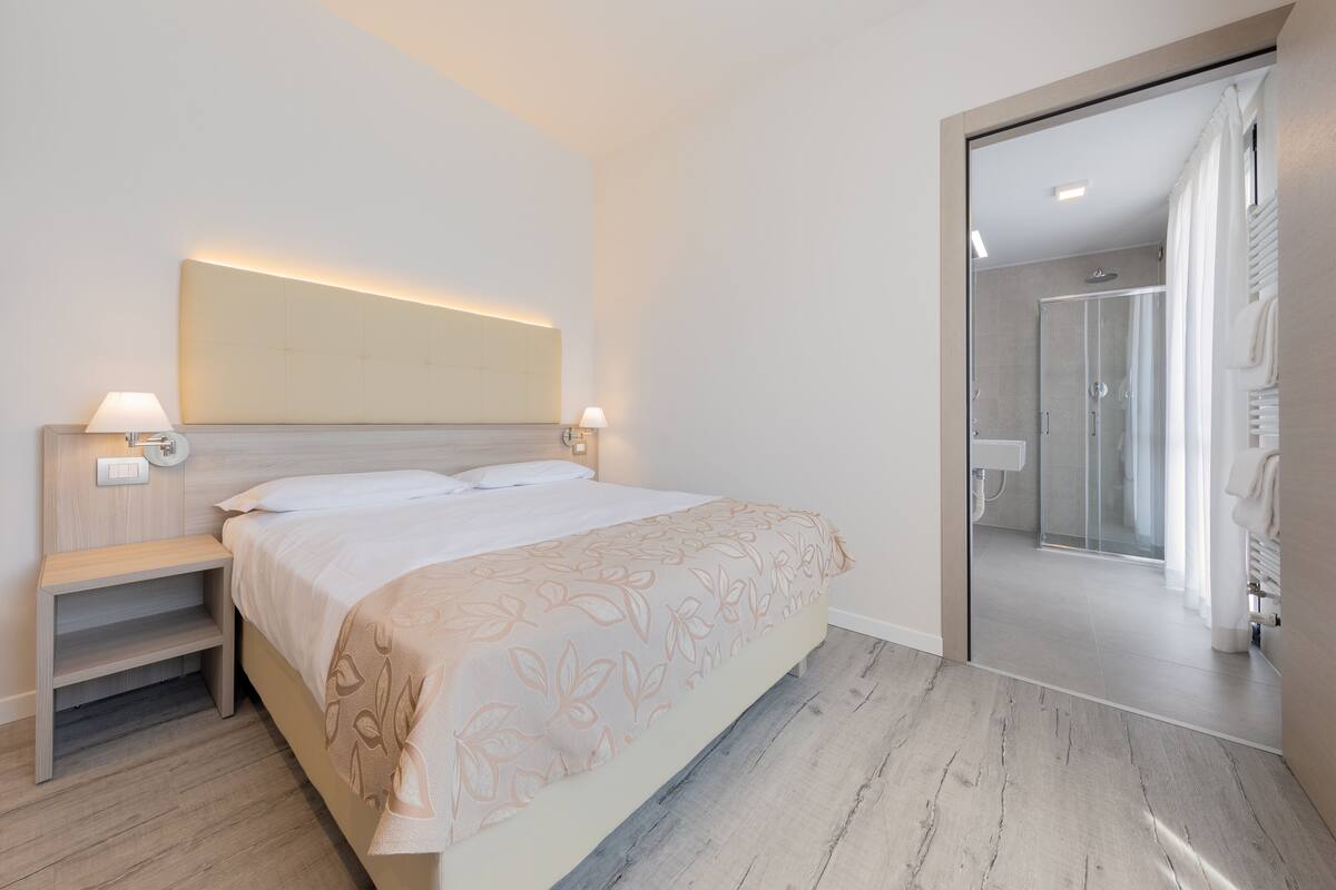 A modern hotel room with a bed, bedside lamps, a large mirror, and a shower area.