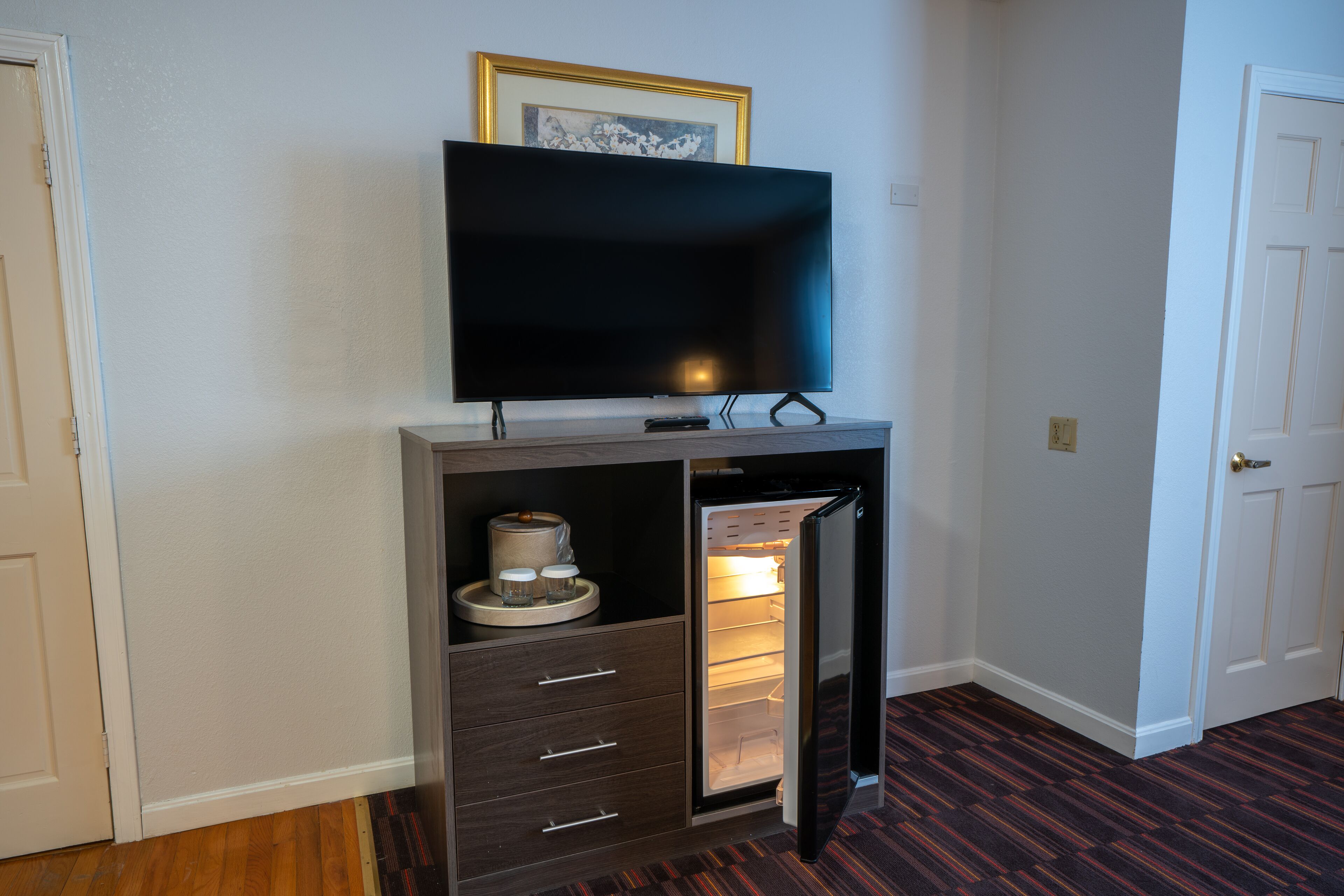 Un espacio habitable compacto con una televisión integrada en un mueble de madera, un refrigerador pequeño y una puerta que da a otra habitación.