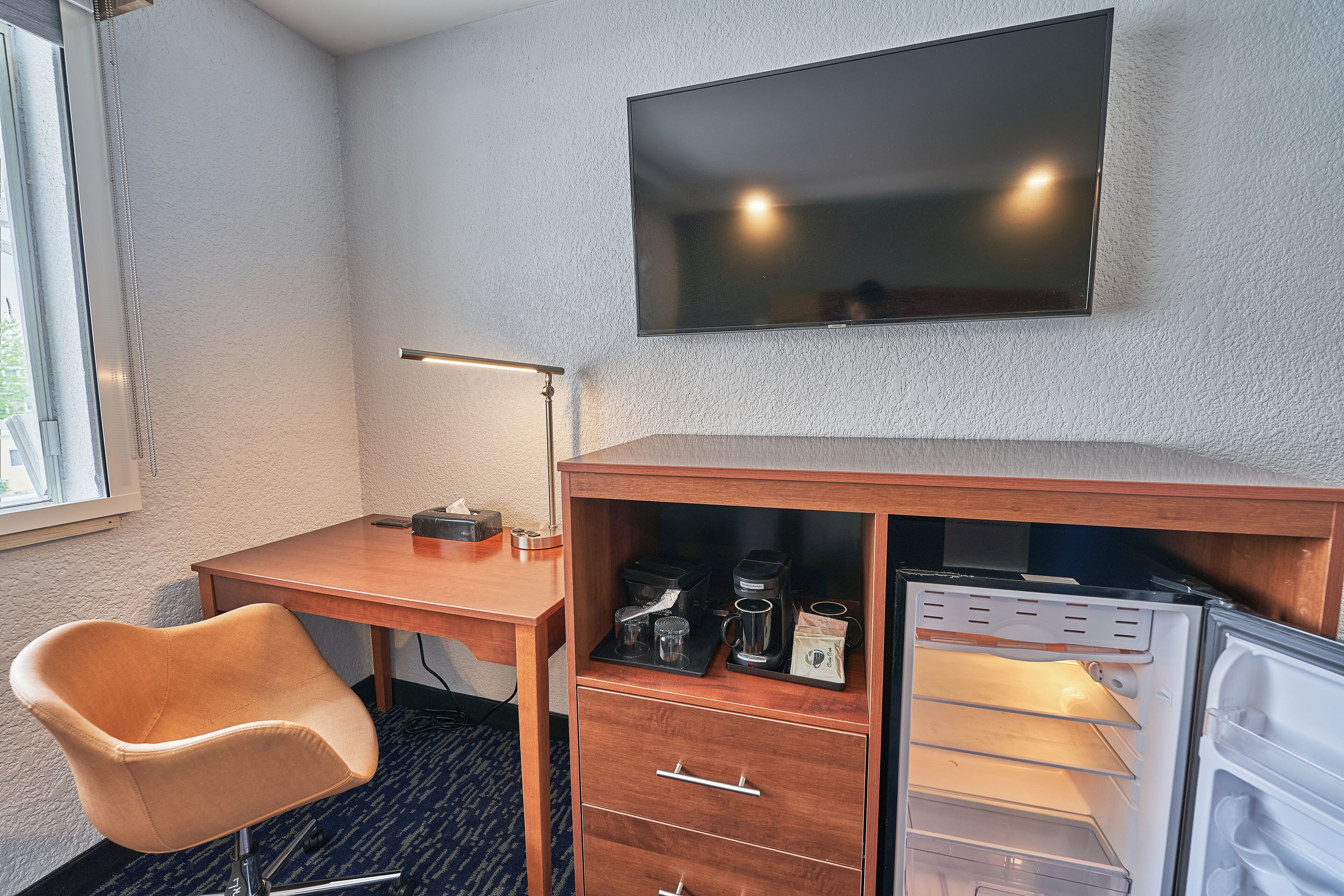 A hotel room with a wooden desk, a chair, a flat-screen TV mounted on the wall, and a minibar with coffee and tea.