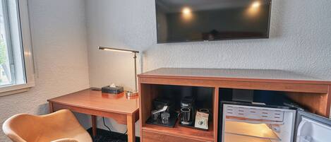 A hotel room with a wooden desk, a chair, a flat-screen TV mounted on the wall, and a minibar with coffee and tea.