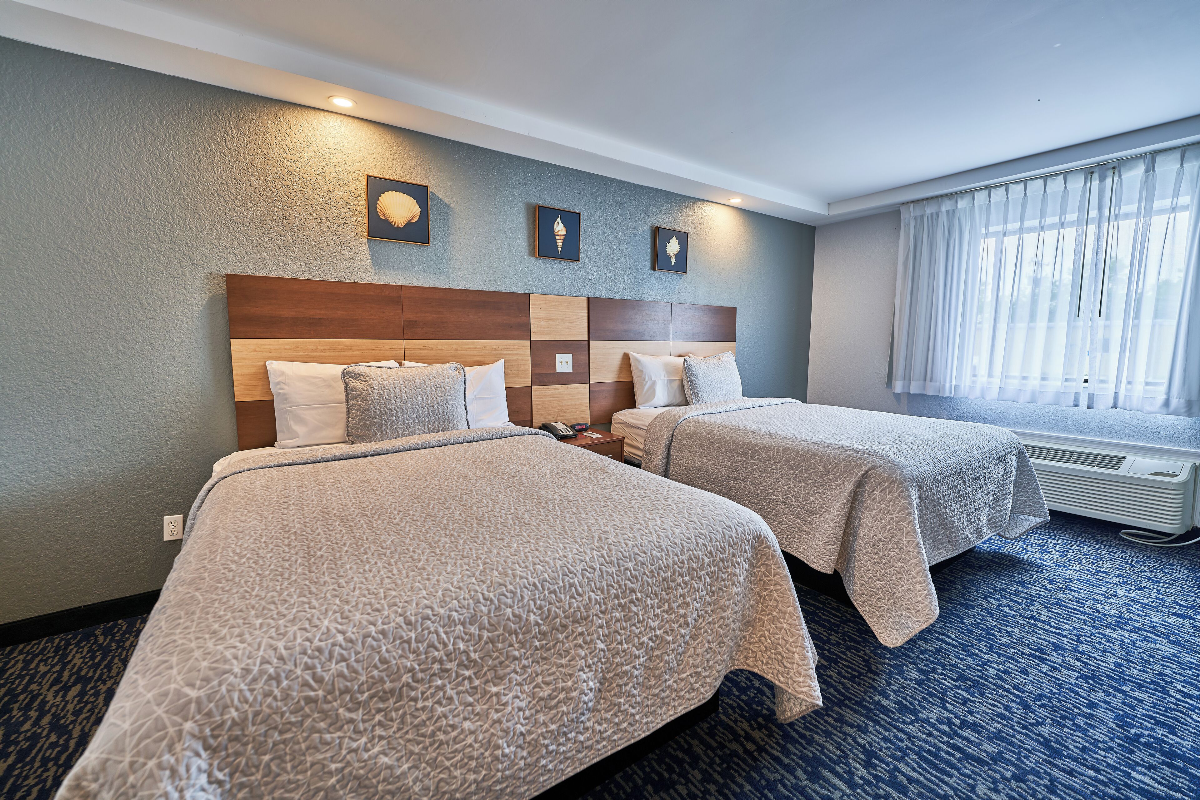 A hotel room with two beds, a large window with sheer curtains, and a wall-mounted air conditioner.