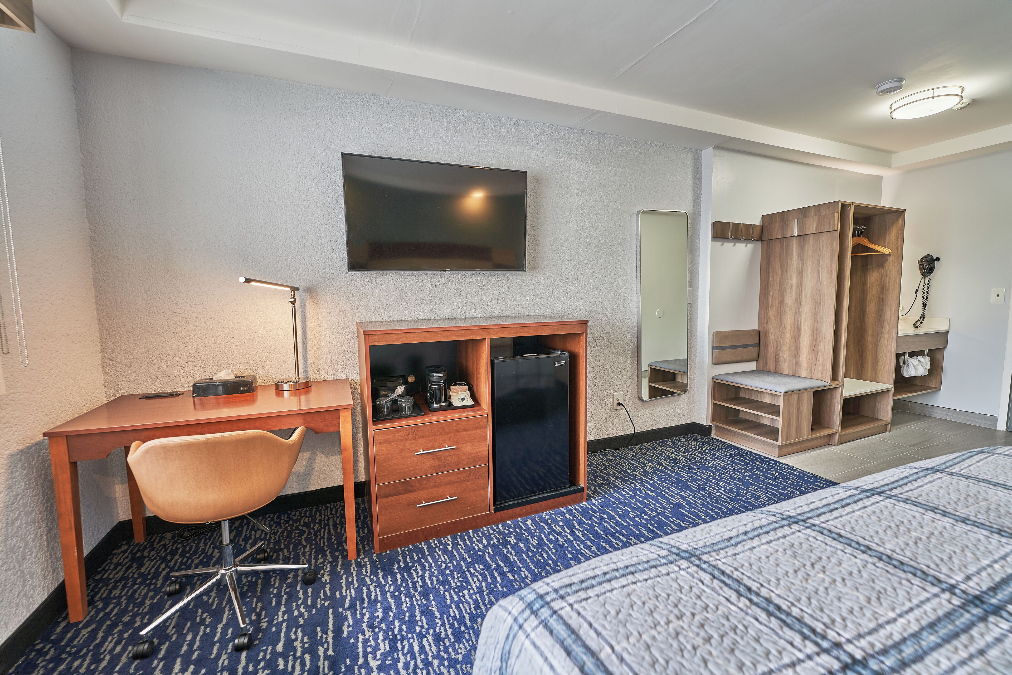 A hotel room with a bed, a desk with a chair, a wardrobe, a television, and a small refrigerator.
