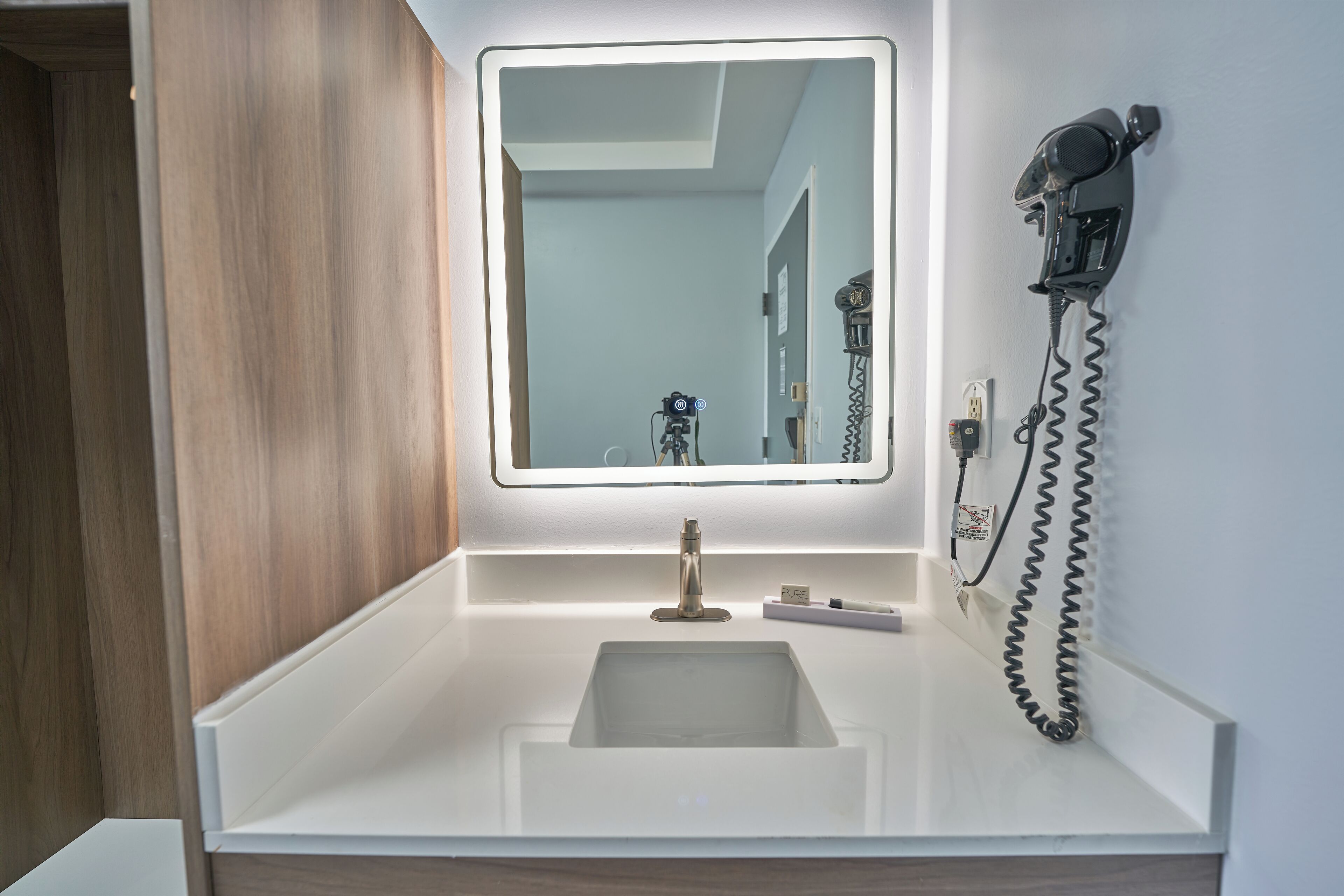 A bathroom with a white sink, a mirror with integrated lighting, and a hairdryer mounted on the wall.