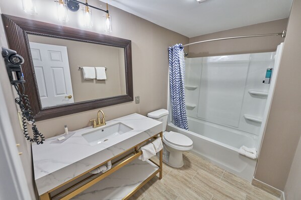 A bathroom with a white sink, gold vanity, a large mirror, a toilet, and a shower with a blue and white striped shower curtain. - Hyannis Plaza Hotel (Hyannis)