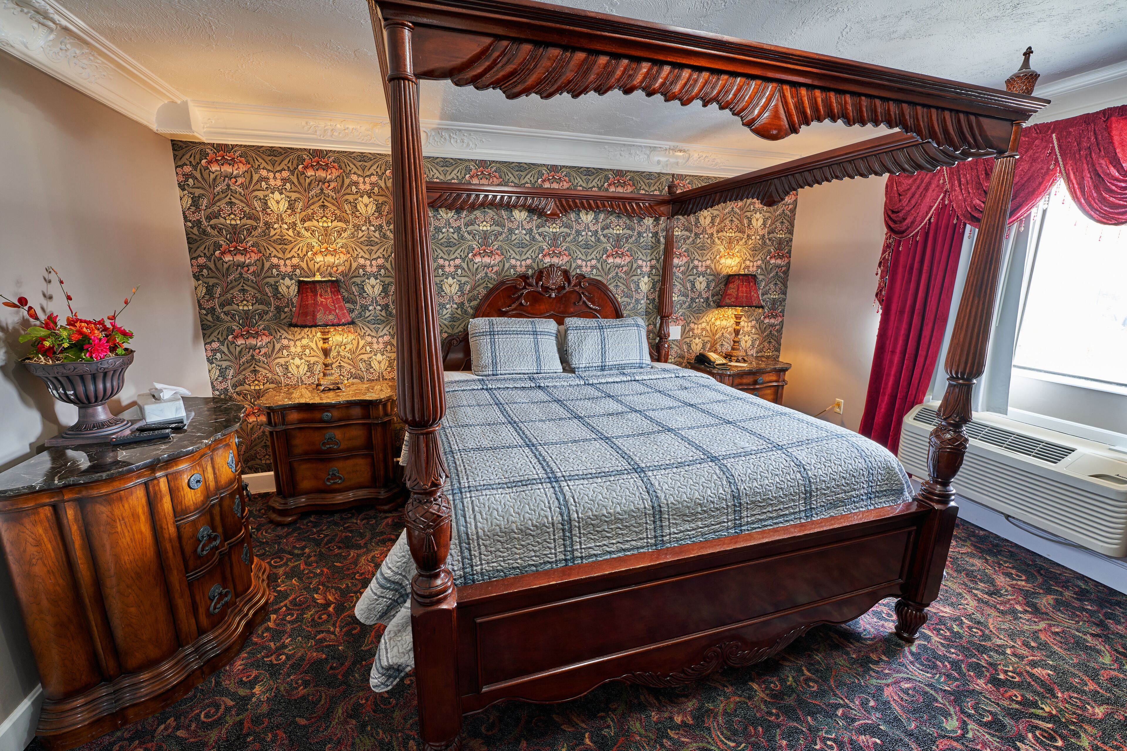 Een hemelbed met een sprei, een houten nachtkastje, een lamp en een vaas met bloemen.