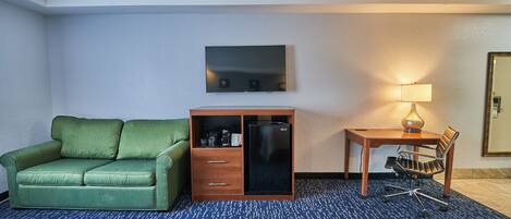 Une chambre d’hôtel avec un canapé vert, un bureau avec une lampe, un téléviseur à écran plat et un mini-bar.