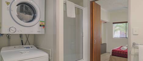 A laundry area with a washing machine and dryer, a glass shower enclosure, and a bedroom with a bed and window.