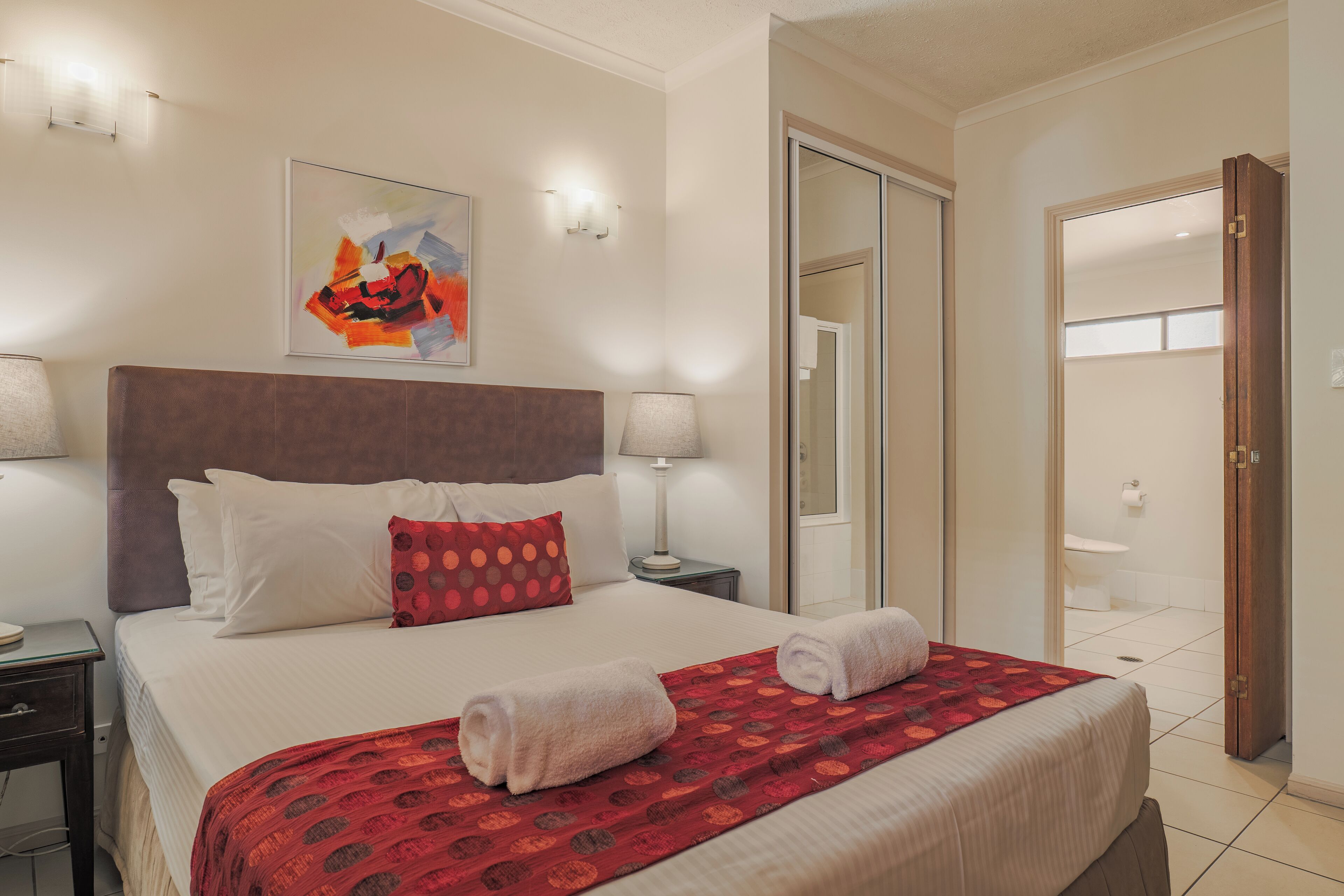 Una habitación de hotel con una cama grande, un cabecero, un cuadro, dos lámparas de noche, una manta roja con estampado y una puerta de madera.
