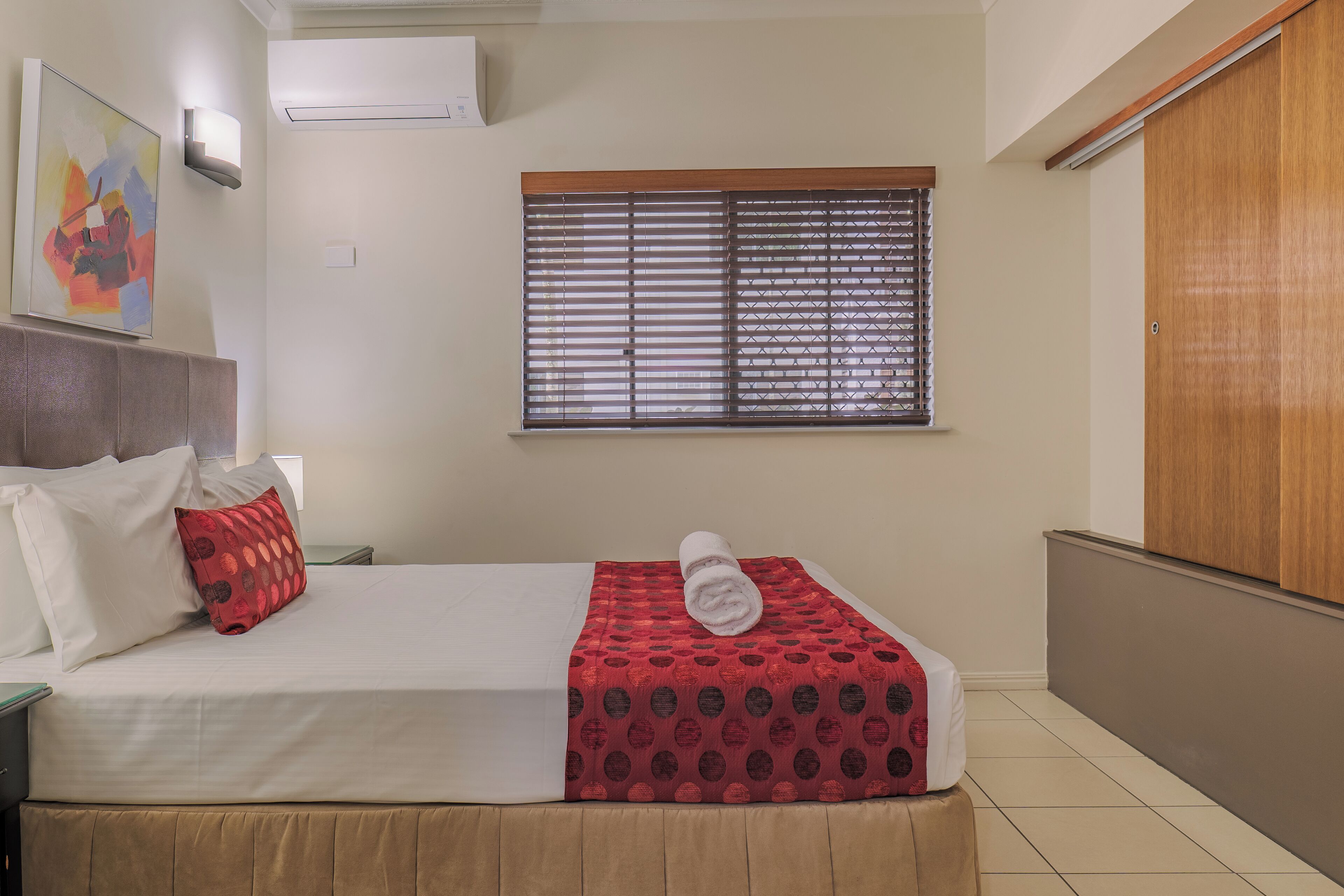 Una cama bien hecha con una colcha roja estampada, una toalla enrollada, una ventana con persianas, un cuadro en la pared y un armario de madera.