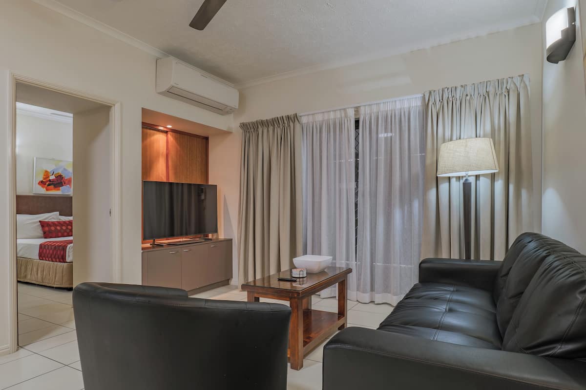 A modern living room with a leather sofa, a wooden coffee table, and a TV. There is a kitchen area with a microwave and cabinets. The room has a large window with curtains.
