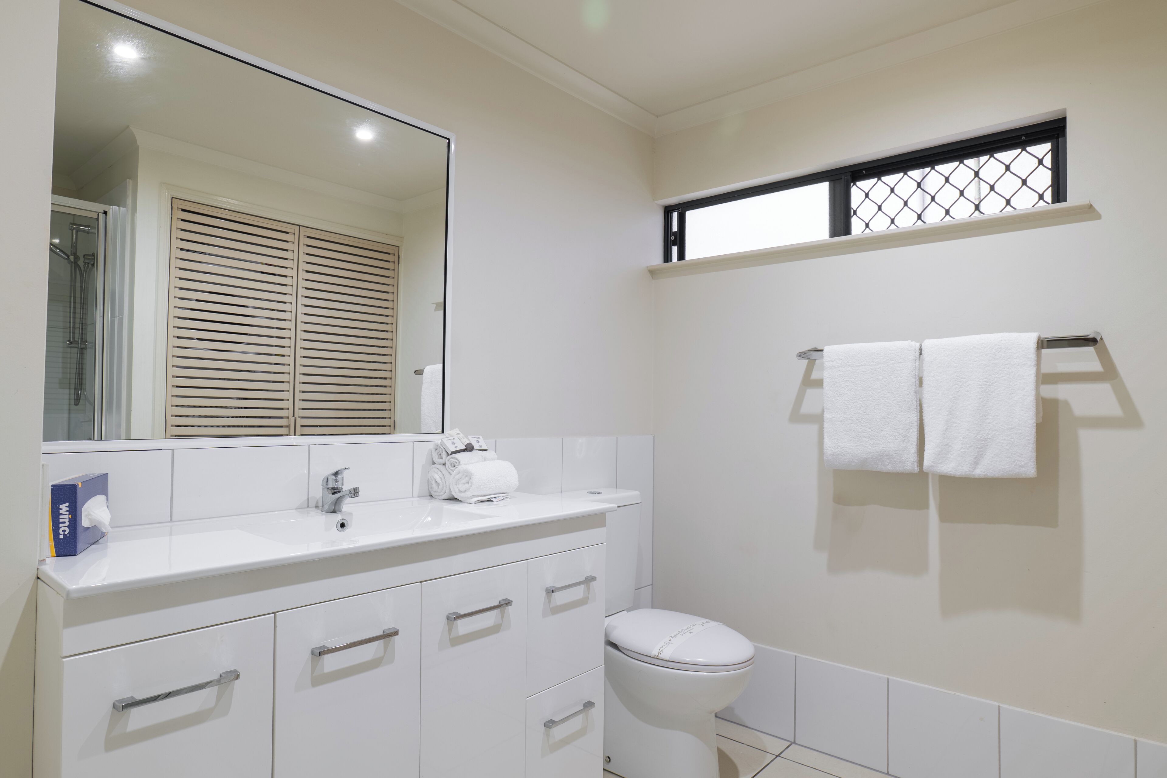 A bathroom with a white sink, mirror, and toilet. There are towels hanging on a rack and a window with a black frame.