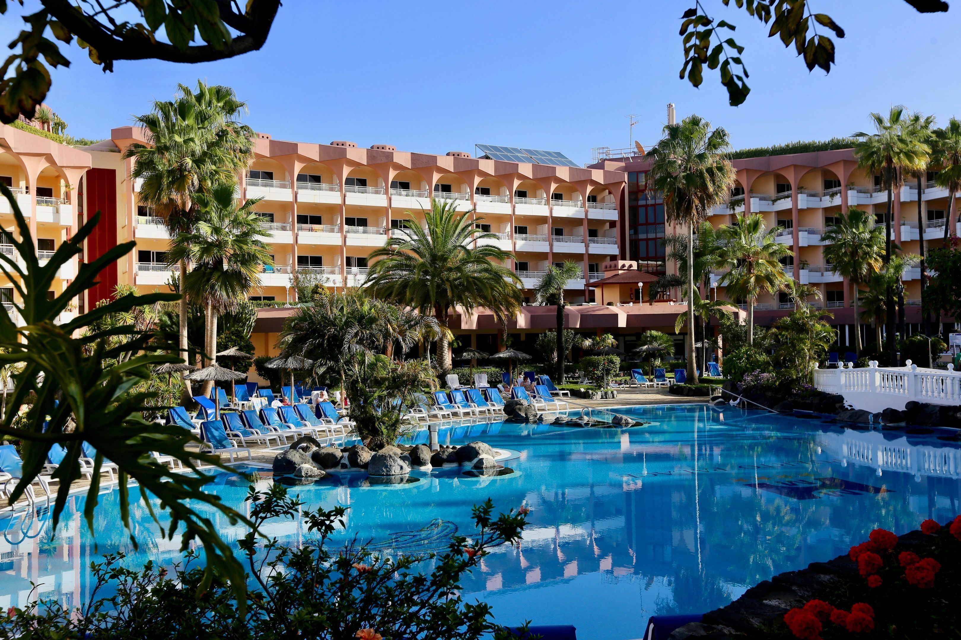 A hotel with a large pool, palm trees, and blue lounge chairs.