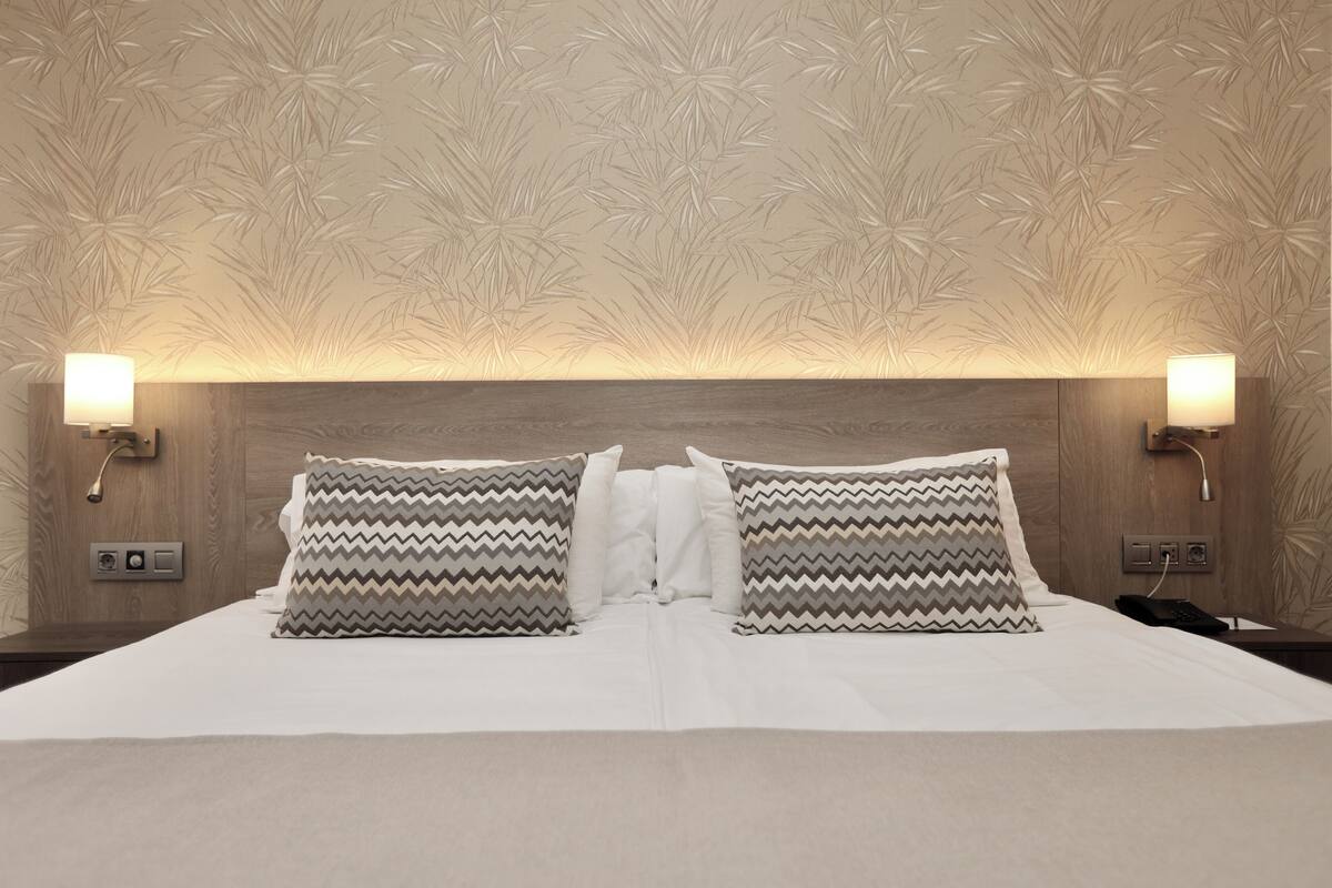 A hotel room with a wooden headboard, two bedside lamps, a bed with white bedding, and patterned pillows.