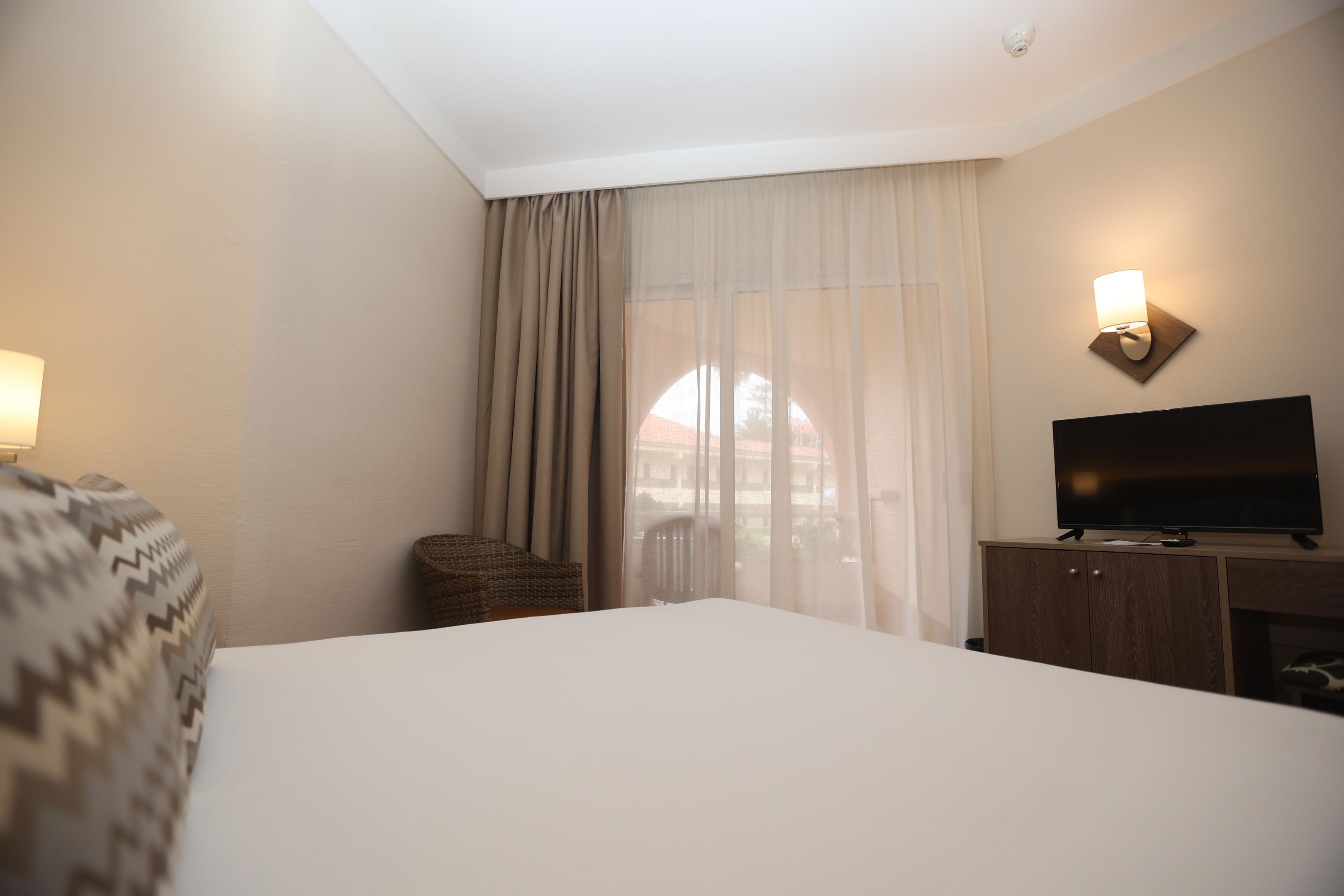 Une chambre d’hôtel avec un grand lit, un téléviseur posé sur une commode en bois, un fauteuil en osier, une fenêtre avec des rideaux légers et un luminaire fixé au mur.
