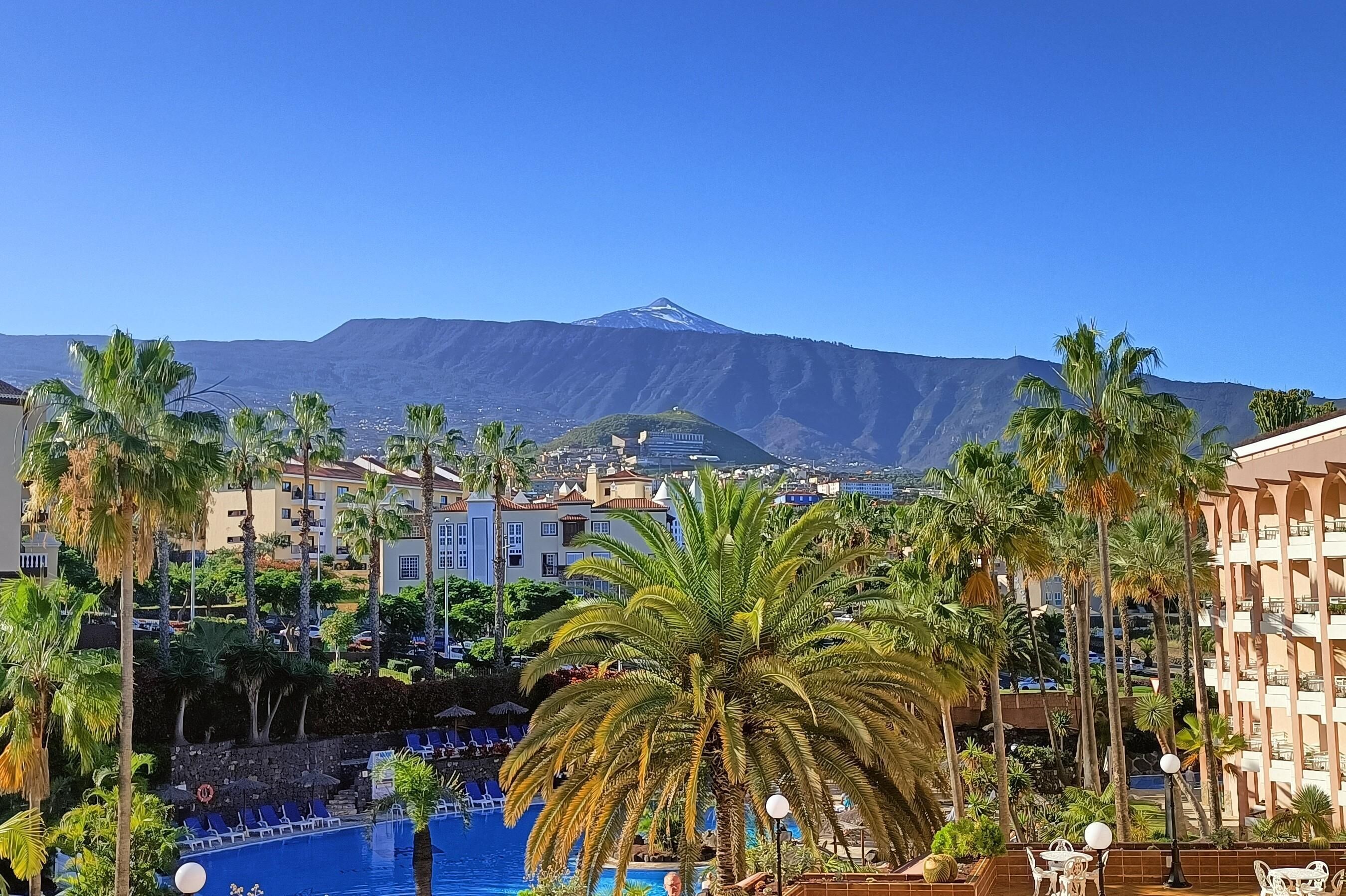 A resort with a pool surrounded by palm trees, a mountain in the background, and hotel buildings.