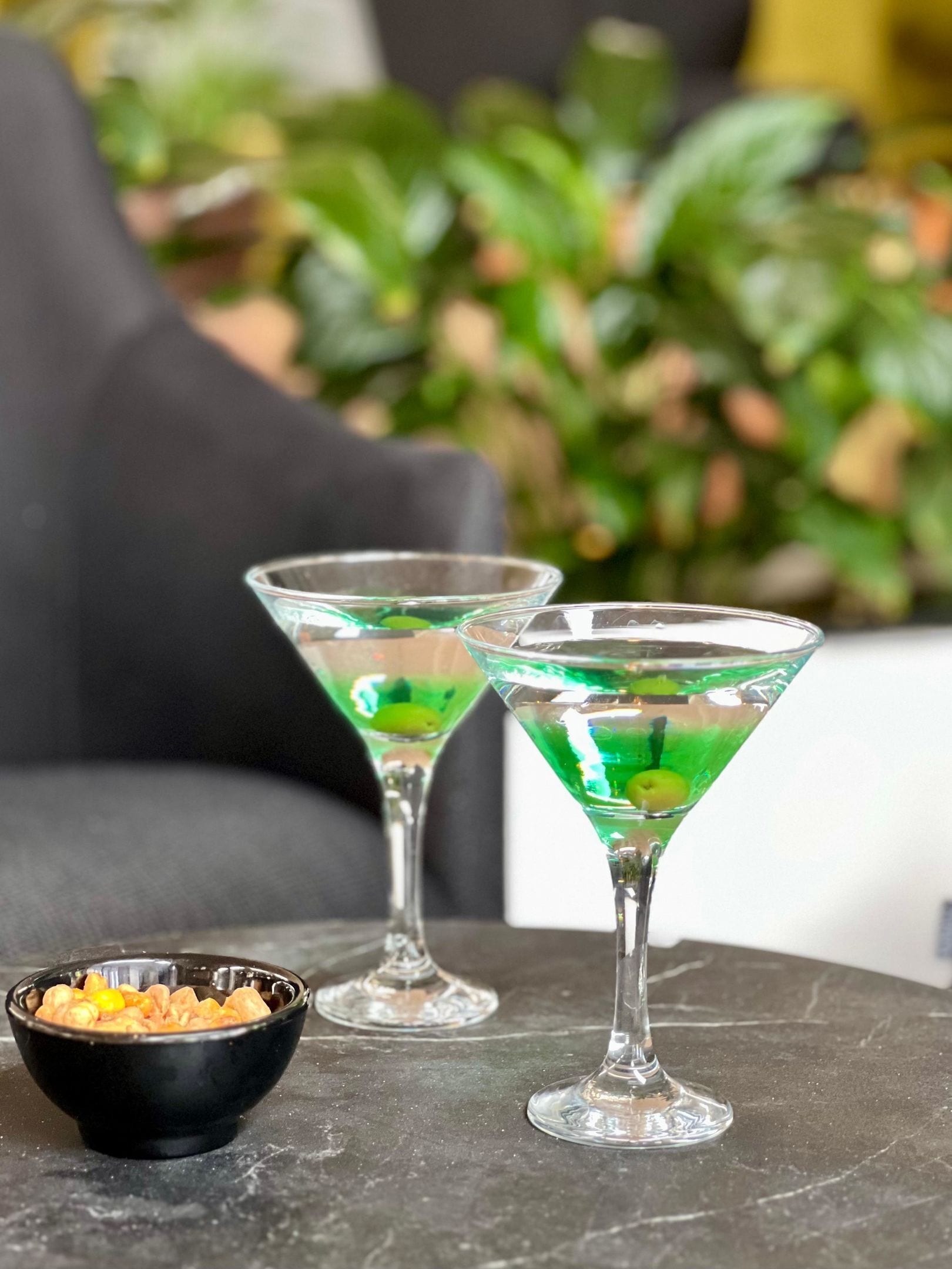 Two martini glasses on a marble table with a black bowl of nuts.