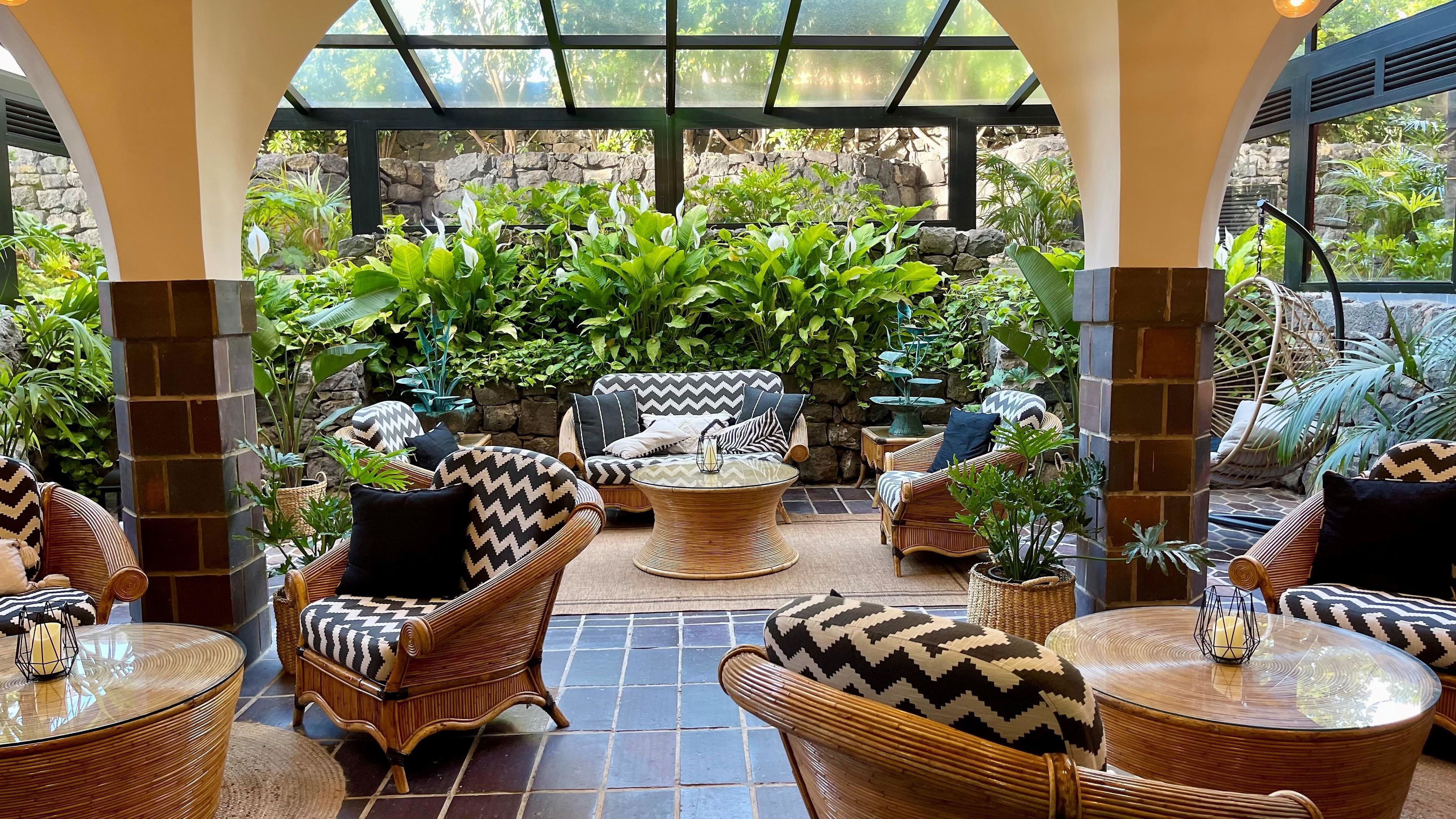 A covered outdoor seating area with wicker furniture, stone walls, and lush greenery.