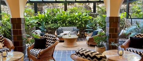 A covered outdoor seating area with wicker furniture, stone walls, and lush greenery.