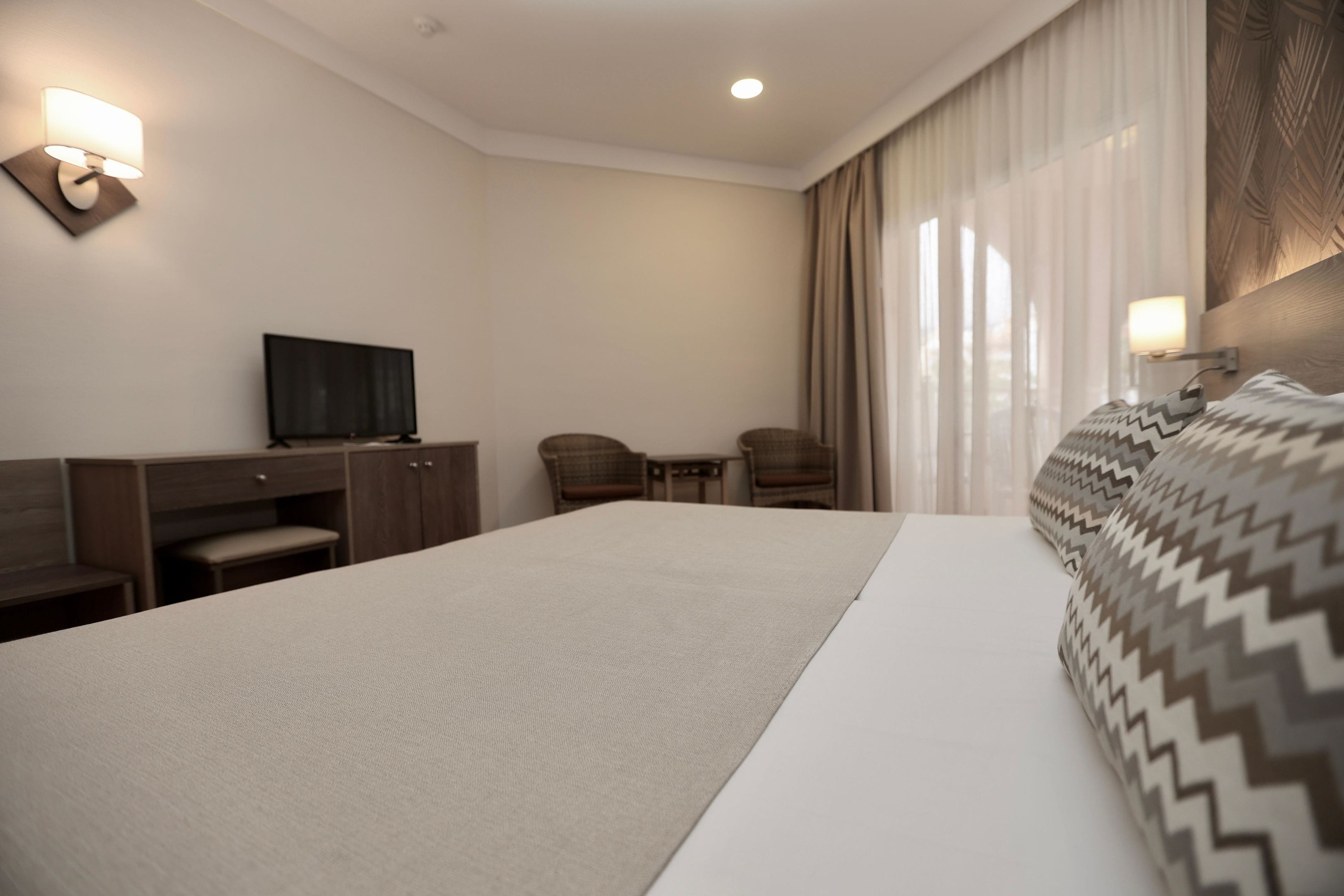 A hotel room with a large bed, a desk with a television, two chairs, and a lamp.