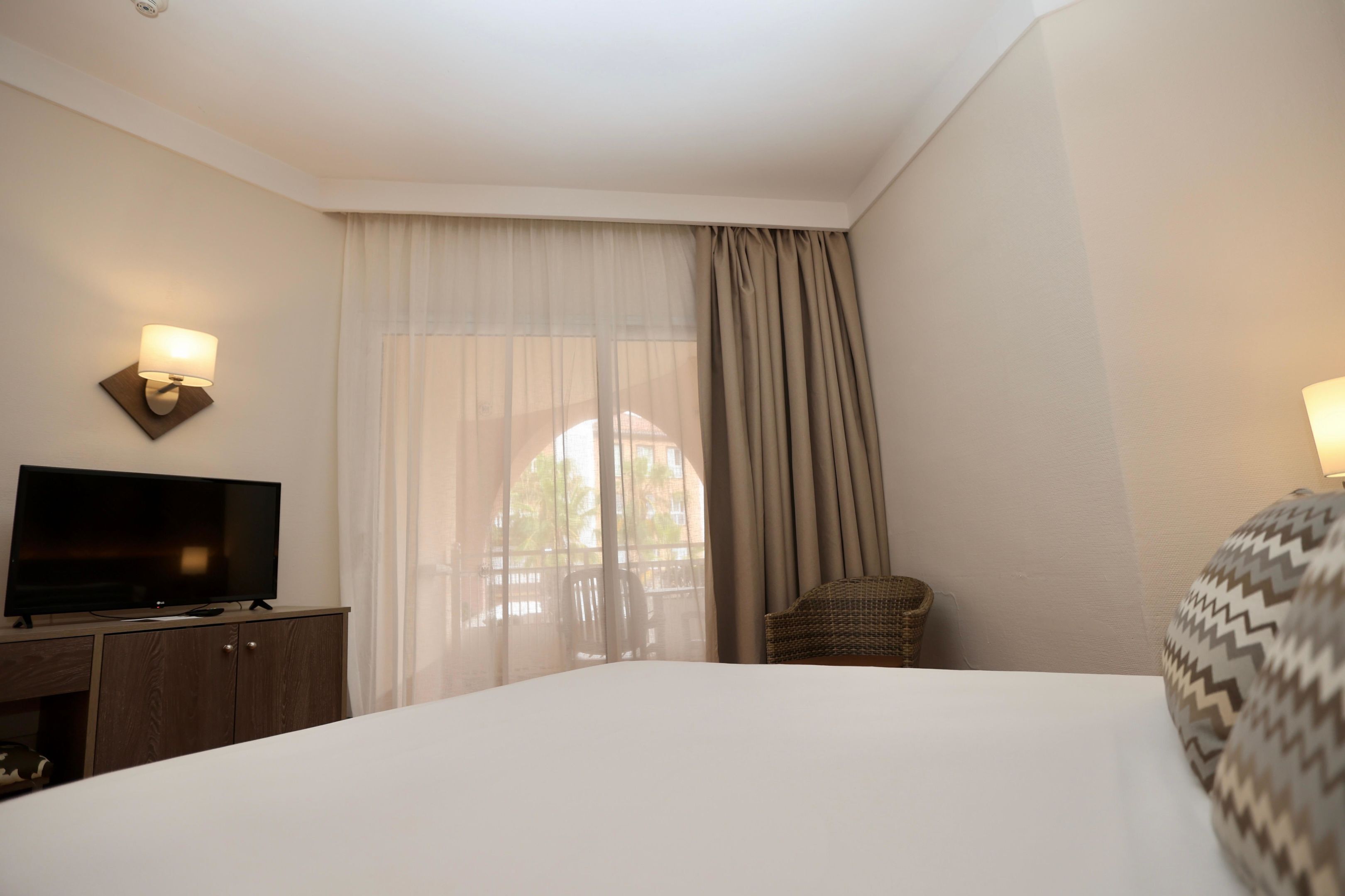 Habitación de hotel con una cama grande, un televisor sobre un mueble de madera, una silla de mimbre y una ventana con cortinas translúcidas.