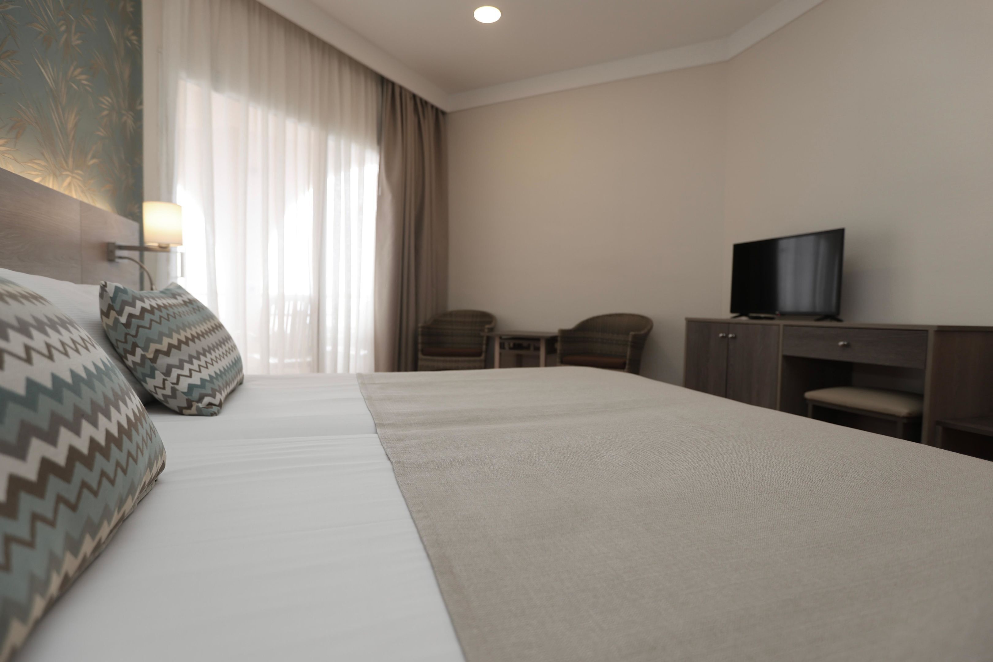Une chambre d’hôtel avec un grand lit, deux fauteuils, un bureau avec une télévision et une fenêtre avec des rideaux.